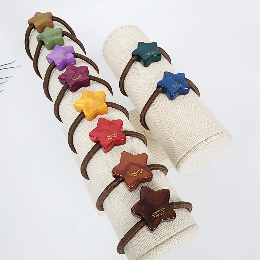 Cellulose Acetate star shaped hair accessory in earth tone and bright colors minimalist geometric ponytail holder for birthday gift. A set of eight star-shaped hair ties on a display stand. The hair ties are made of different colors, including green, purple, red, orange, yellow, and brown. Each hair tie has the words 'CREATION FRANCE' printed on it.