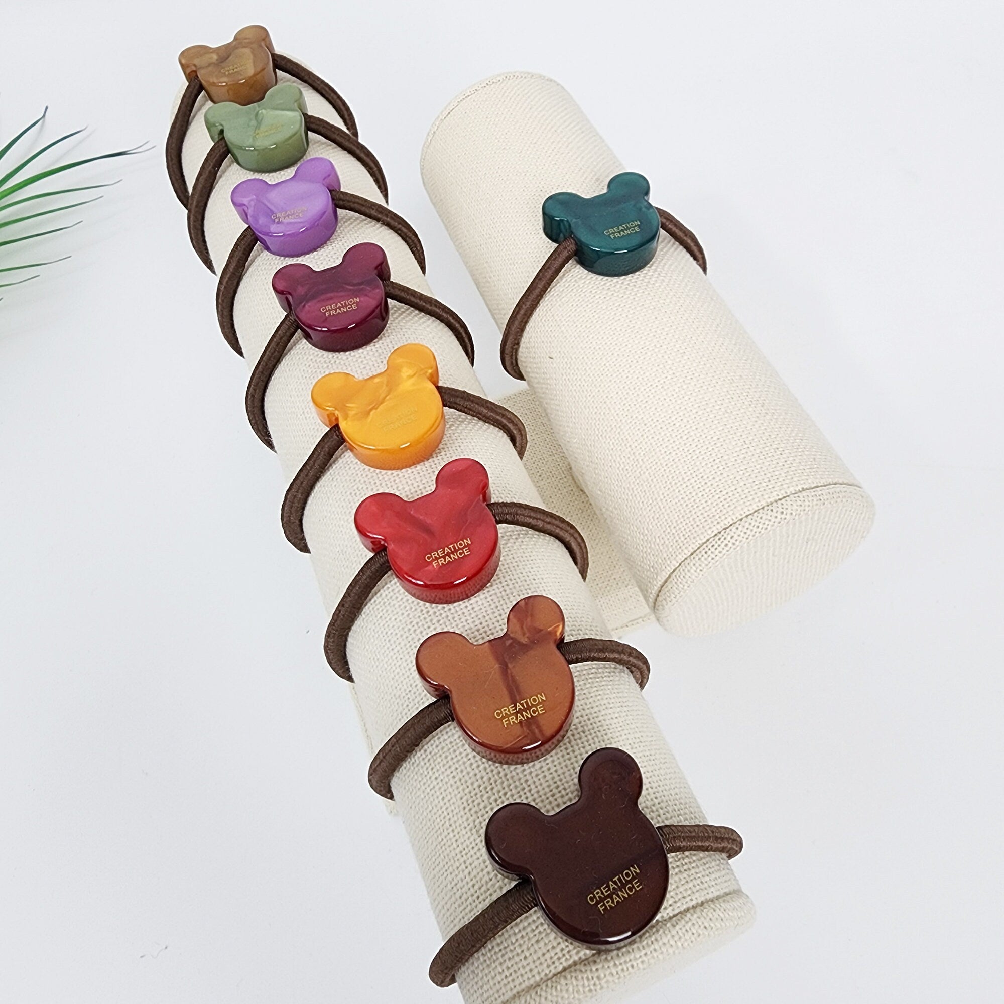 Cellulose acetate mouse shaped hair tie with velvet aurora finish, cute animal charm ponytail holder with durable elastic band for women and girls. Eight brown hair ties with colorful Mickey Mouse shaped resin embellishments. The embellishments have the words 'CREATION FRANCE' printed on them. The hair ties are displayed on a white fabric covered cylinder.