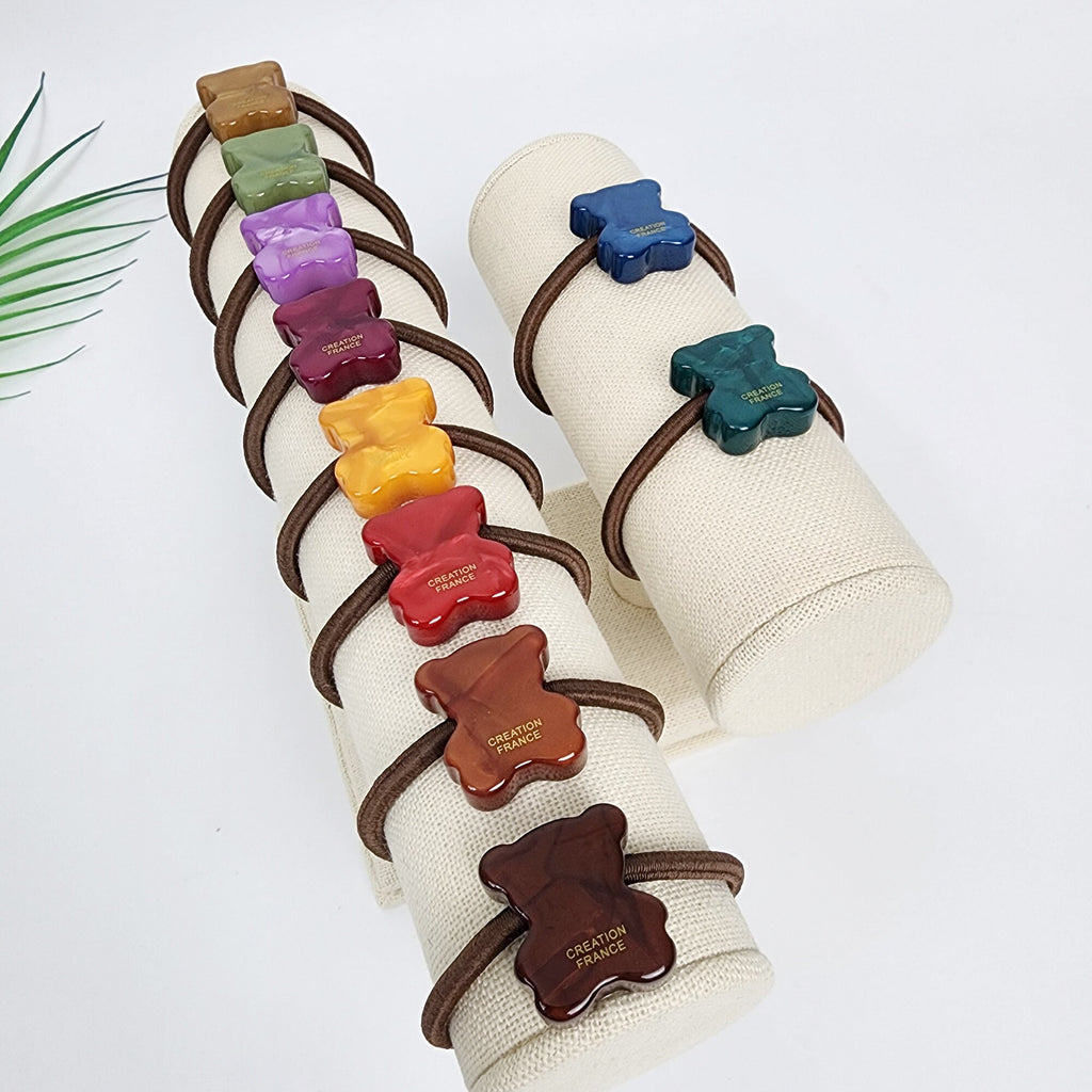 Cellulose acetate teddy bear hair tie with glossy aurora finish, cute minimalist animal ponytail holder for women and girls. A set of ten colorful hair ties with a bear design. The hair ties are arranged on two white fabric covered display stands. The bear designs are made of resin and have the words 'Creation France' printed on them. The colors of the hair ties are brown, red, yellow, purple, green, blue, and orange.