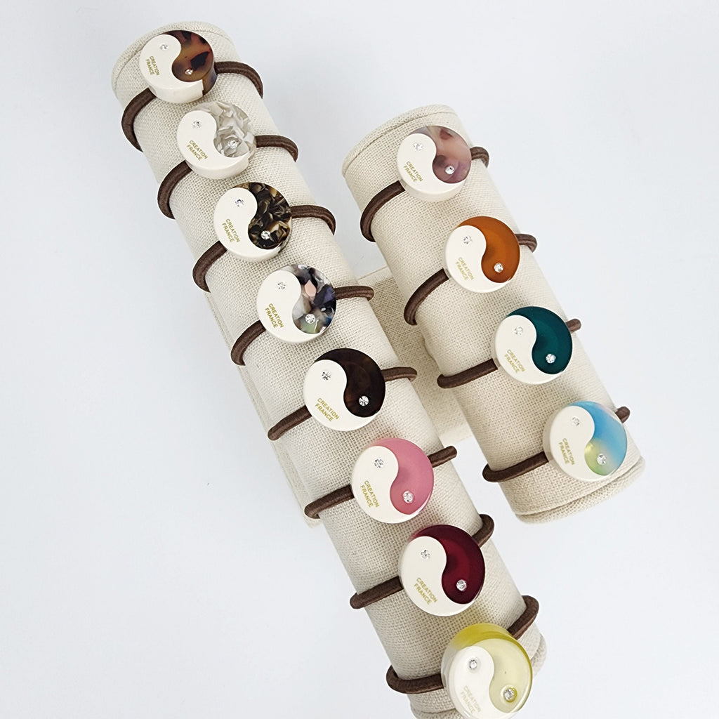 Cellulose acetate round hair tie with crystal center in white and pastel colors. Circle ponytail holder for women. Korean minimalist hair accessory. Two white stands with brown elastic bands holding various color yin yang hair ties. The hair ties have a gold inscription that reads 'CREATOR PEACE'.