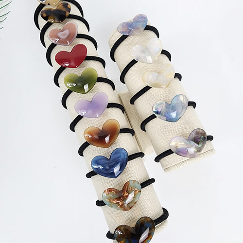 Glossy cellulose acetate heart shaped hair tie in tortoise shell and pastel colors, convex love charm ponytail holder with durable black elastic band for women and girls. Two sets of hair ties with heart-shaped charms. The charms are made of different colors and patterns, including brown, red, green, purple, blue, and white. The hair ties are displayed on two white stands.