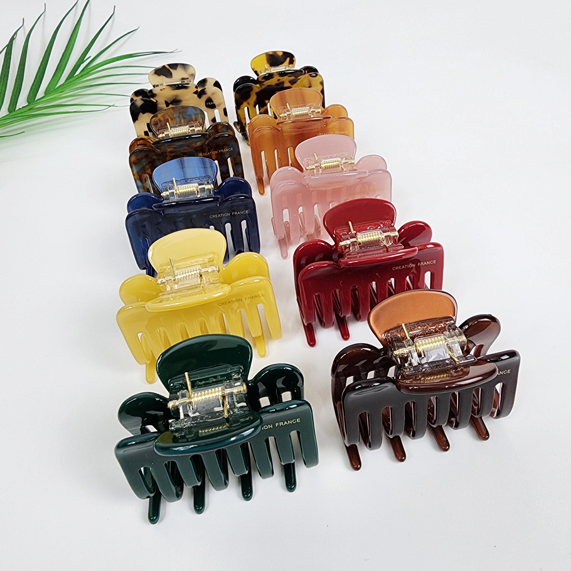 Cellulose acetate zigzag hair claw clip in tortoise shell and solid colors. Small 2.2 inch glossy nonslip jaw clamp for women creating half updo hairstyles. A set of ten hair claw clips in various colors including brown, yellow, green, red, orange, blue, and pink. The clips are made of plastic and have a textured finish. Each clip has a gold metal spring and the words 'Creation France' printed on the side.