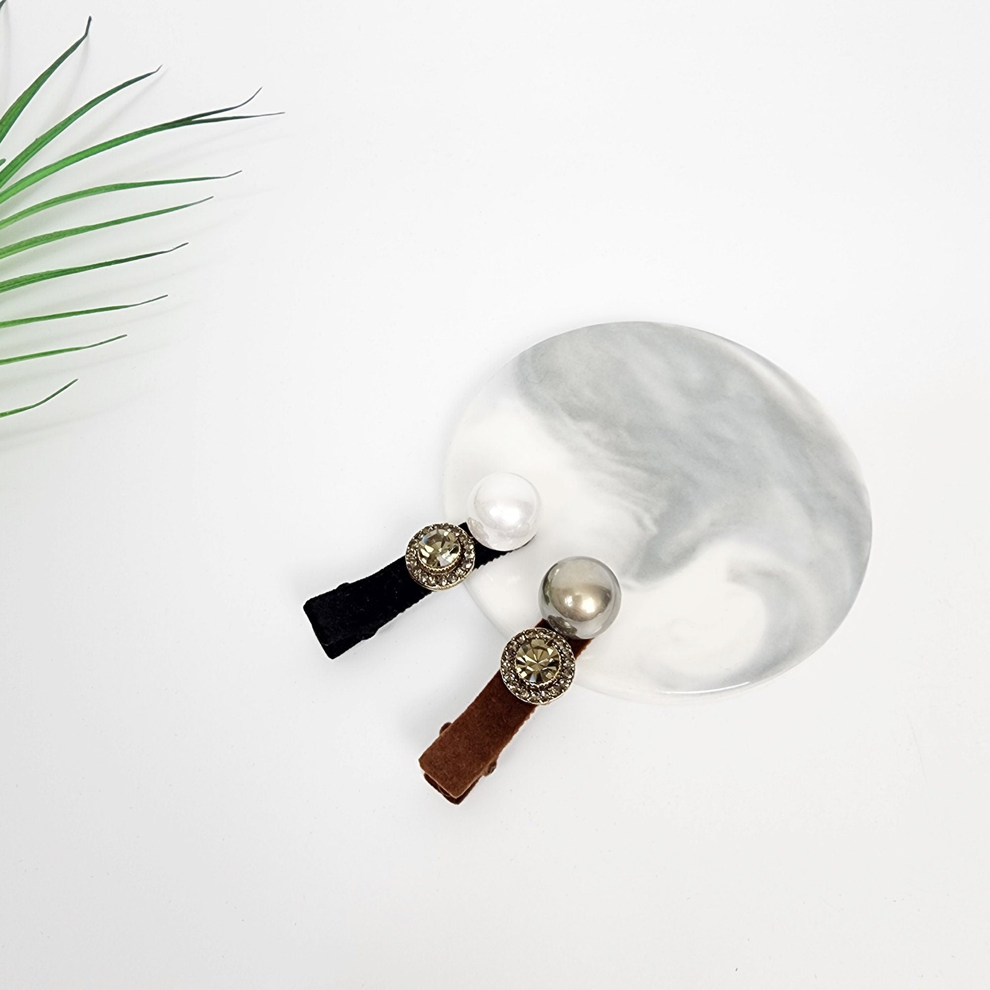 Mini velvet alligator clip with faux pearl and rhinestone button. 2-inch side bangs barrette for elegant styling. Two hair clips with brown and black velvet ribbon and a gold rhinestone embellishment. The clips are on a white marble surface with two pearl-like beads.