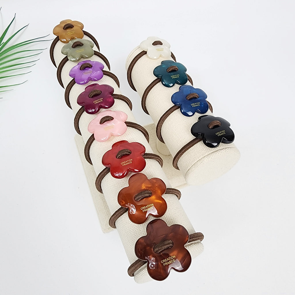 Cellulose acetate flower hair tie with stitch detail. Convex floral ponytail holder in 12 colors. Korean retro hair accessory for women. A display of 12 hair scrunchies with flower-shaped buttons. The scrunchies are arranged in two columns on a white fabric stand. The buttons are made of different colors including red, pink, brown, green, blue, black, and white. Each button has the words 'CREATION FRANCE' printed on it.