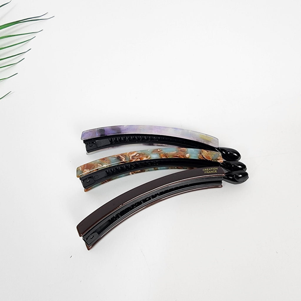 Cellulose acetate slim banana clip with inner teeth mechanism for fine hair and half updo styling. Three hair clips with a curved shape and a black base. The clips are decorated with a marbled pattern in shades of purple, brown, and blue. The bottom clip has the text 'CREATION FRANCE' printed on it.