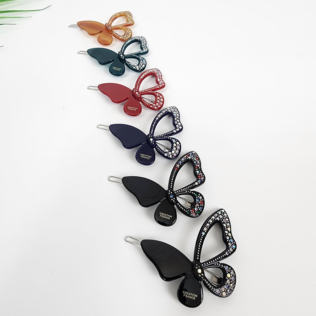 Butterfly shaped cellulose acetate hair clip with crystal rhinestone details. Sparkly snap barrette for side bangs, available in black, red, and green. Six butterfly hair clips with rhinestones. The clips are in various colors including orange, green, red, navy blue, black, and black. The clips have the words 'CREATION FRANCE' printed on them.