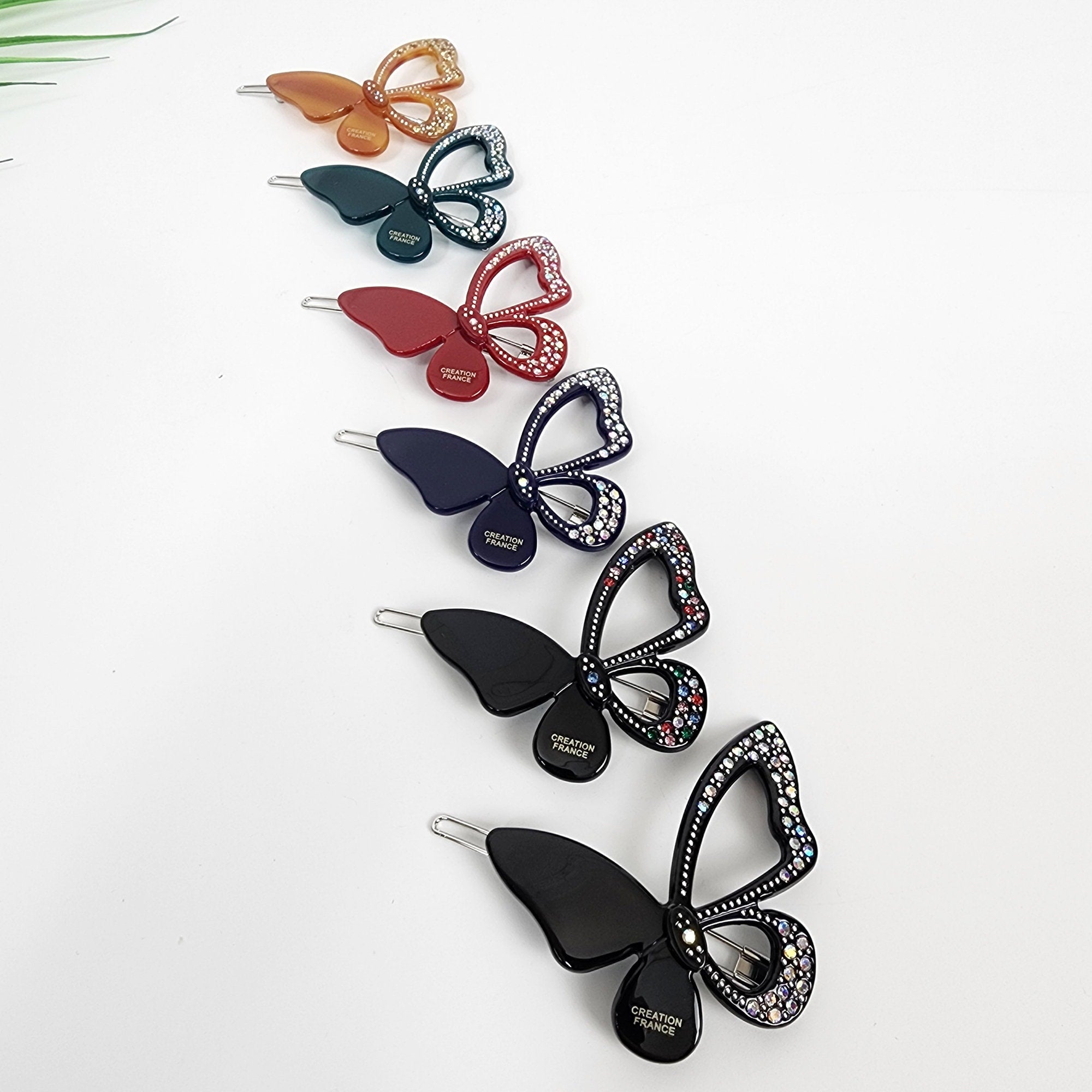 Butterfly shaped cellulose acetate hair clip with crystal rhinestone details. Sparkly snap barrette for side bangs, available in black, red, and green. Six butterfly hair clips with rhinestones. The clips are in various colors including orange, green, red, navy blue, black, and black. The clips have the words 'CREATION FRANCE' printed on them.