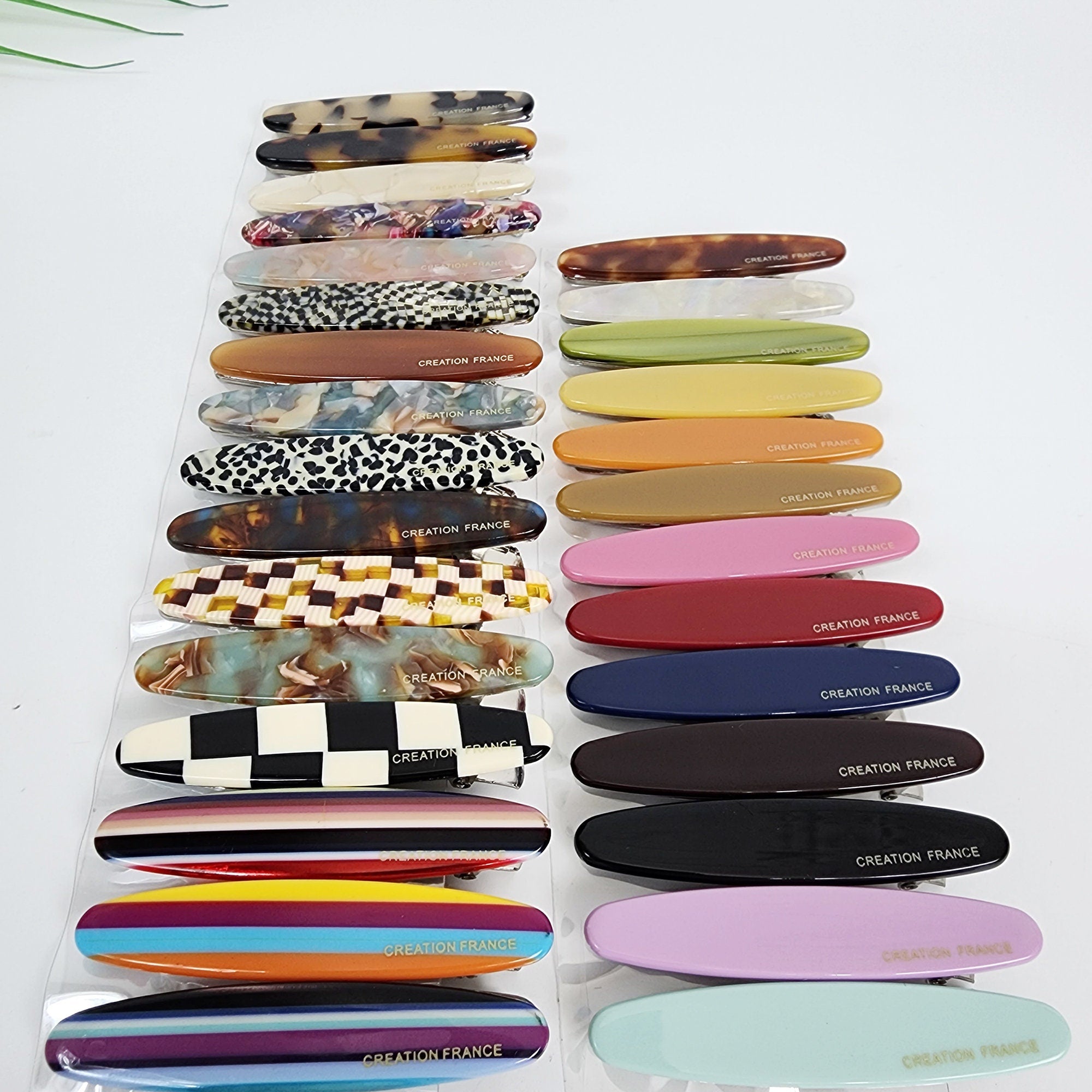 Cellulose acetate slim oval barrette, 2.5 inch alligator beak clip for side bangs, colorful minimalist hair accessories for women. A collection of 18 colorful plastic hair clips. The clips are all oval shaped and have a silver metal clasp. The clips have various patterns including checkered, striped, floral, and abstract designs. The clips have the text 'CREATION FRANCE' printed on them.