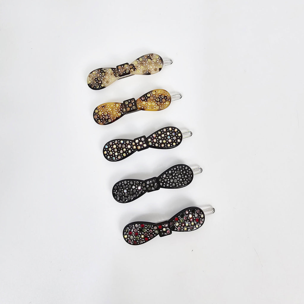 Cellulose acetate bow hair clips with rhinestone embellishments. Small 2 inch snap barrettes in tortoise shell and black, perfect for bangs and side hair styling. Five hair clips in the shape of bows with rhinestones. The bows are in various colors including brown, black, and multi-colored. The clips are arranged vertically on a white background.