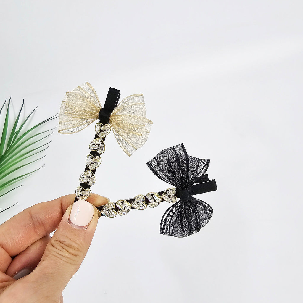 Gold and black mesh bow hair clips featuring a row of heart-shaped cubic zirconia crystals on a metal beak clip. Elegant 3.1 inch side slide barrettes with sparkling jewel details and sheer ribbon accents, perfect for formal styling or bangs management. Two hair clips with black ribbon bows and rhinestone accents. One bow is beige and the other is black.
