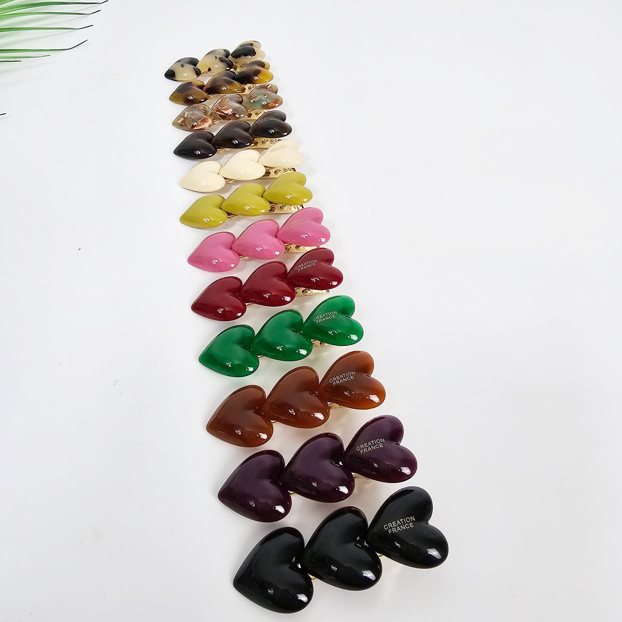 Cellulose acetate triple heart hair clip in pink and tortoise shell, mini alligator clip for bangs, cute side hair pin for women. A row of double heart-shaped hair clips in various colors including black, purple, brown, green, red, pink, yellow, white, and tortoise shell. Each clip has the words 'Creation France' printed on the back.