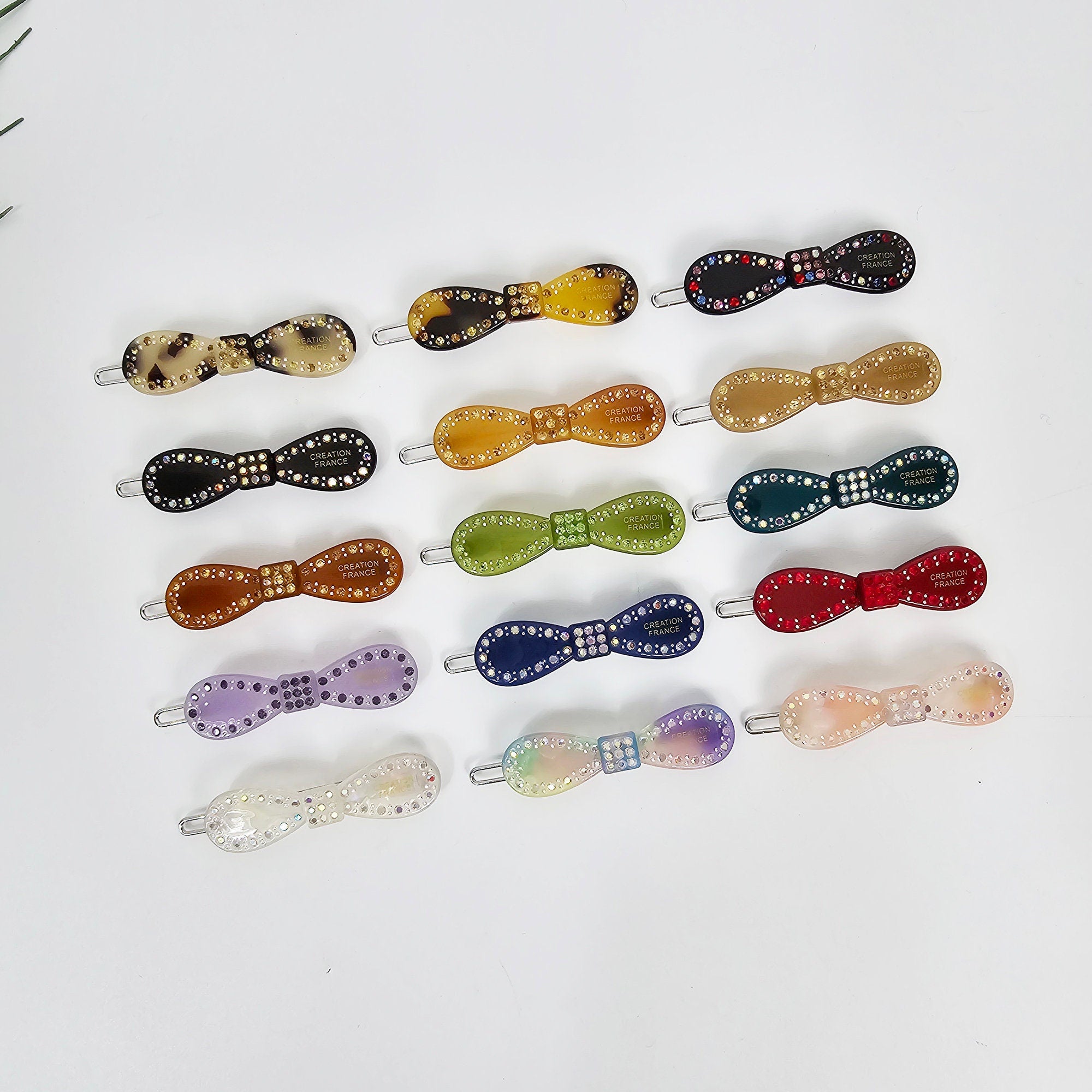 Cellulose acetate bow shaped hair barrettes with crystal accents. Small 2.0 inch tortoise shell and solid color snap clips, perfect for holding bangs or side hair with a secure click-clack mechanism.  A collection of 15 hair clips with a bow design. The clips are made of various colors including brown, tan, green, blue, purple, red, and white. Each clip has a small rhinestone bow and the words 'Creation France' printed on the back.