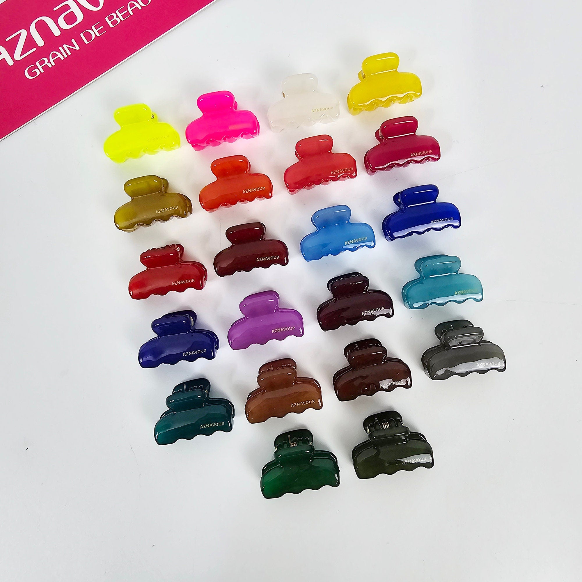 Cellulose acetate mini rectangular hair claw clips in 22 vibrant colors. Small 1 inch non-slip jaw clips for bangs or half-up styles, featuring a durable spring and glossy finish. A collection of 18 colorful plastic hair clips. The clips are all the same shape and size, with a curved, claw-like design. The colors include yellow, pink, red, orange, brown, blue, purple, green, and gray. Each clip has the word 'AZANOUR' printed on it in gold lettering.