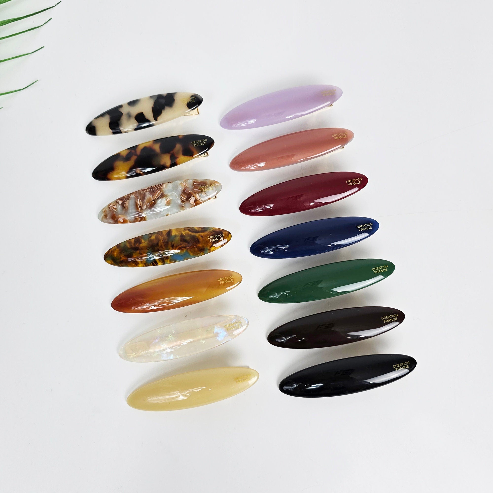Oval shaped cellulose acetate alligator hair clips in purple, tortoise shell, and solid colors. Sleek 7cm flat beak barrette for women's side bangs styling. Korean minimalist hair accessory. A collection of fourteen assorted color and patterned hair clips. The clips are all oval shaped and have a gold logo that says 'CREATION FRANCE'.