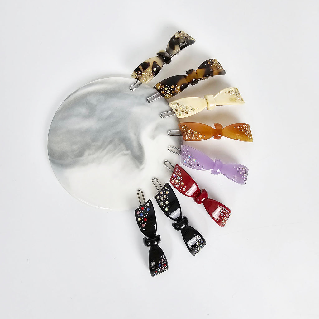 Cellulose acetate bow snap clip with rhinestone crystals, tortoise shell and colorful patterns, small hair barrette for bangs and side hair. Eight hair clips with a bow shape. The clips are made of plastic and have a variety of colors including black, red, purple, brown, beige, and tortoise shell. Each clip has a small amount of rhinestones on the bow.