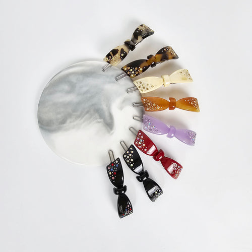 Cellulose acetate bow snap clip with rhinestone crystals, tortoise shell and colorful patterns, small hair barrette for bangs and side hair. Eight hair clips with a bow shape. The clips are made of plastic and have a variety of colors including black, red, purple, brown, beige, and tortoise shell. Each clip has a small amount of rhinestones on the bow.