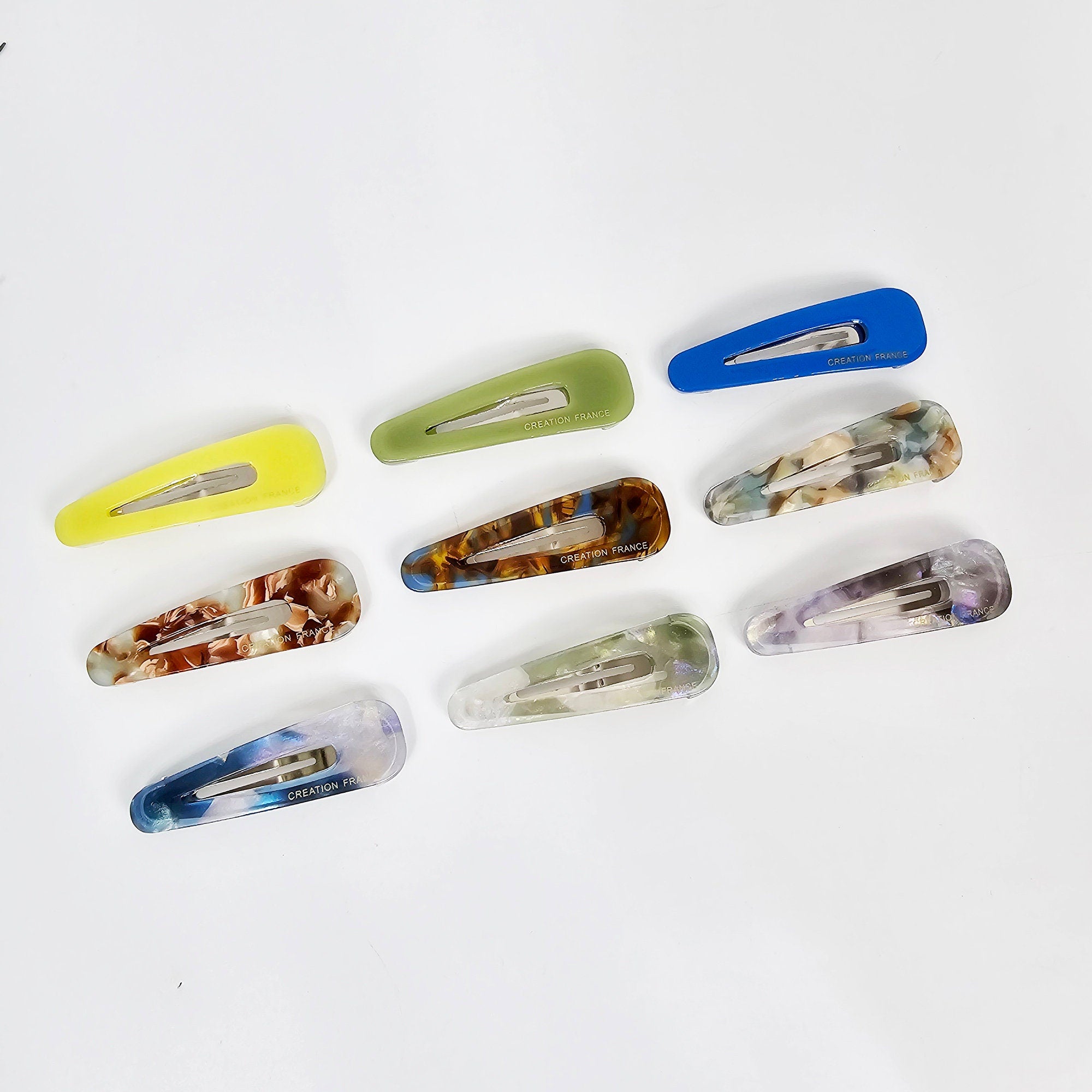Alt Text Cellulose acetate triangle snap barrettes featuring tortoise shell patterns and solid pastel colors for minimalist women styling. Nine assorted plastic hair clips with a variety of colors and patterns. The clips are all rectangular and have a slight curve. The colors include green, blue, brown, and white. The patterns include floral, marble, and abstract designs. The clips have the words 'CREATION FRANCE' printed on them.