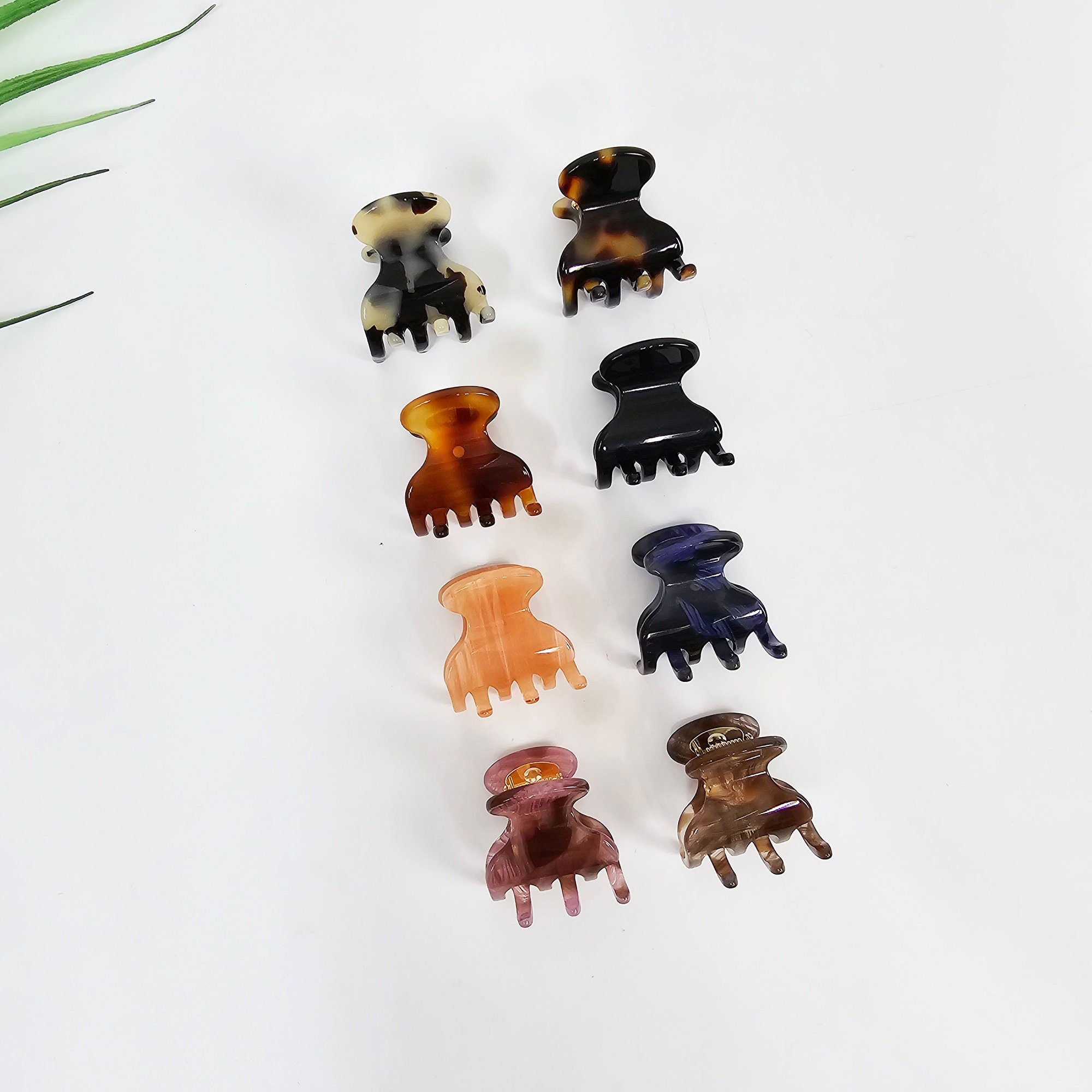 Cellulose acetate mini hair claw clip in tortoise shell and peach, small 1.2 inch jaw clamp for half up hairstyle. Seven small plastic hair clips in various colors and patterns. The clips are arranged in two rows. The top row has three clips: one brown and black, one brown and white, and one black. The bottom row has four clips: one peach, one purple, one dark blue, and one brown and white.