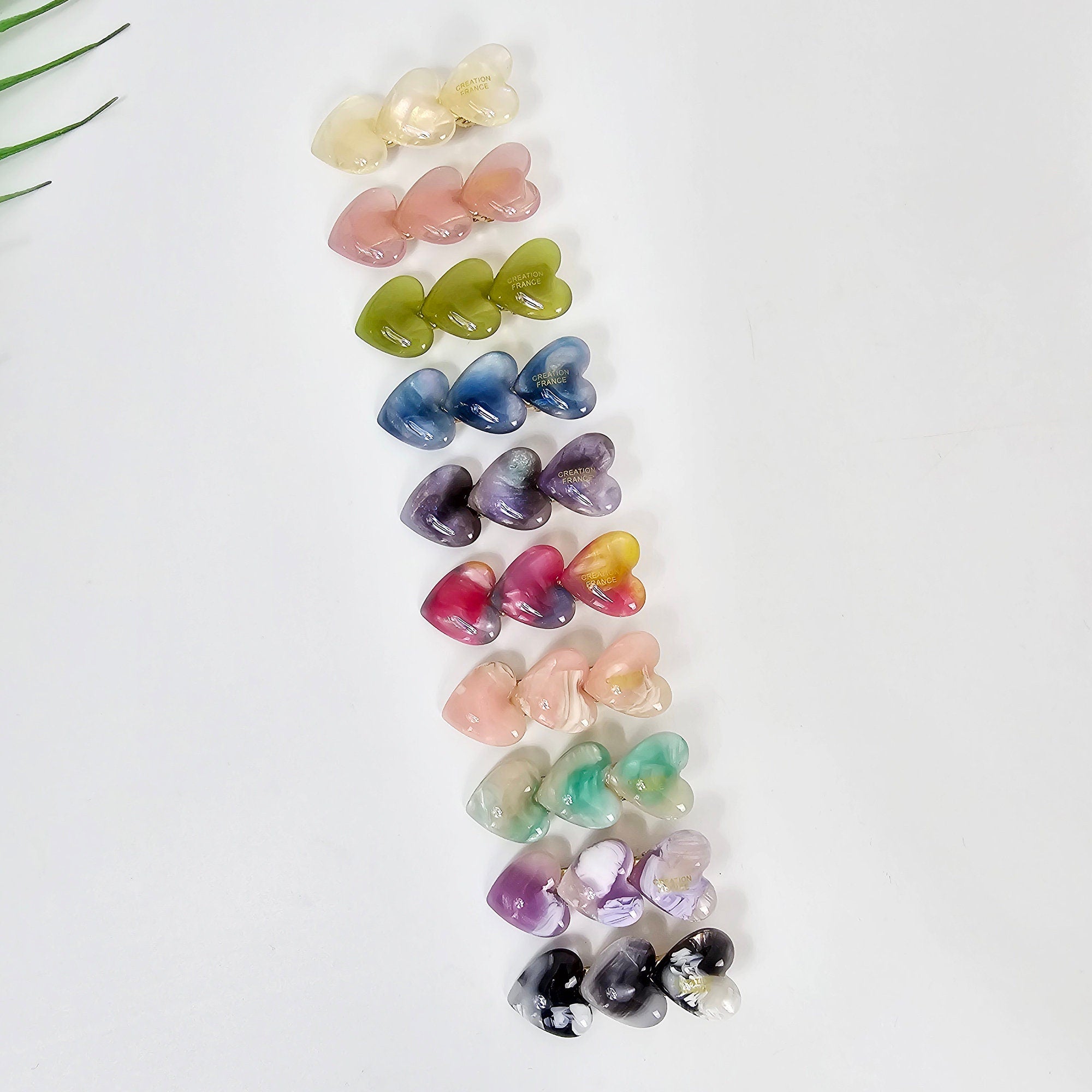 Cellulose acetate hair clip with three small hearts in a row. Mini alligator barrette in tortoise and pastel colors for side bangs styling. A set of 14 heart-shaped hair clips in various colors, including white, pink, green, blue, purple, and black. The clips are made of resin and have a glossy finish. Each clip has a unique swirl pattern and a gold accent.