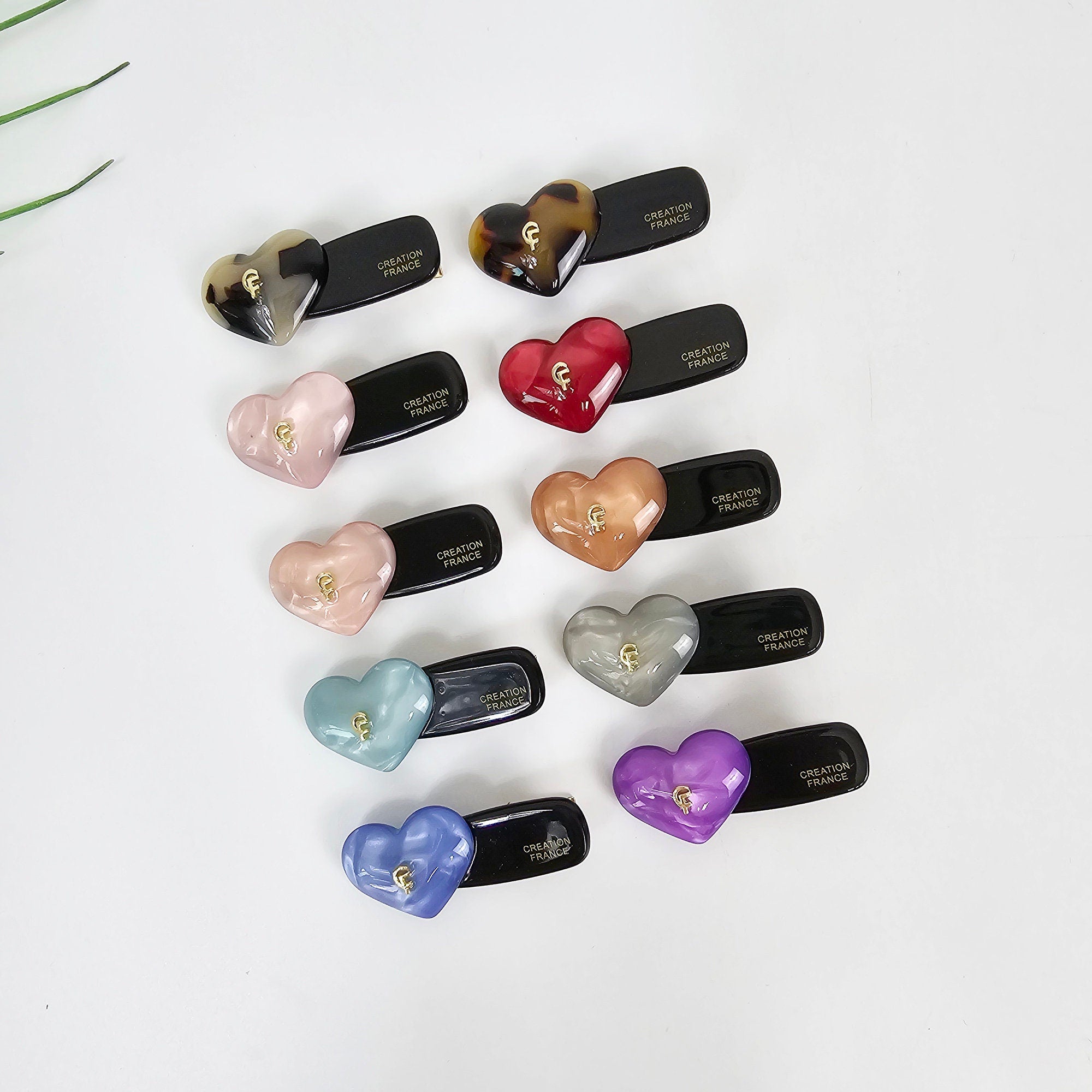 Cellulose acetate mini heart hair clip with gold logo. Small alligator barrette in tortoise and pastel colors for girls and side bangs. A collection of twelve heart-shaped hair clips in various colors, including brown, red, pink, light pink, blue, light blue, purple, and gray. Each clip has a black base with the words 'CREATION FRANCE' printed on it.