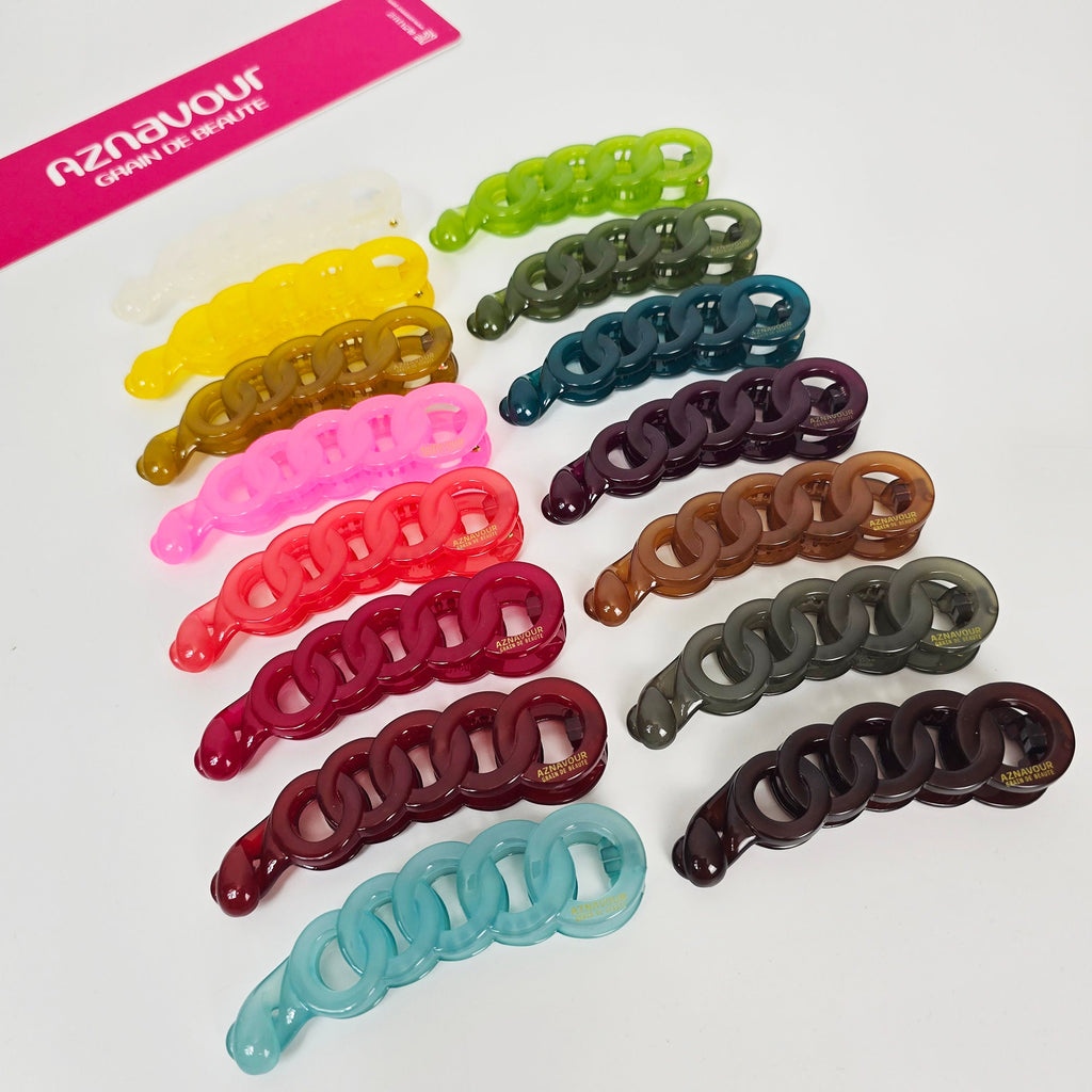 Cellulose acetate banana clip with a chain link design in white. 3.9 inch vertical ponytail holder with inner teeth for secure grip on thick hair.  A set of ten plastic hair clips in various colors, including green, brown, pink, red, and blue. The clips are shaped like chains with interlocking links. The clips are all from the brand 'Aznavour Grain de Beaute'.