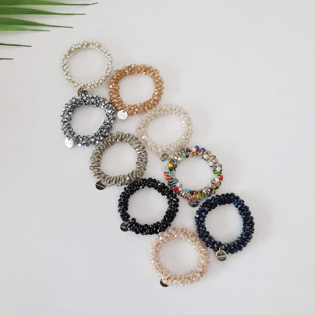 Sparkling glass bead hair tie bracelet with charm, handmade crystal elastic ponytail holder for women. A set of eight beaded hair ties in various colors, including silver, gold, black, and multi-colored. The hair ties are made of small beads and are coiled into a spiral shape. Each hair tie has a small metal tag attached to it.