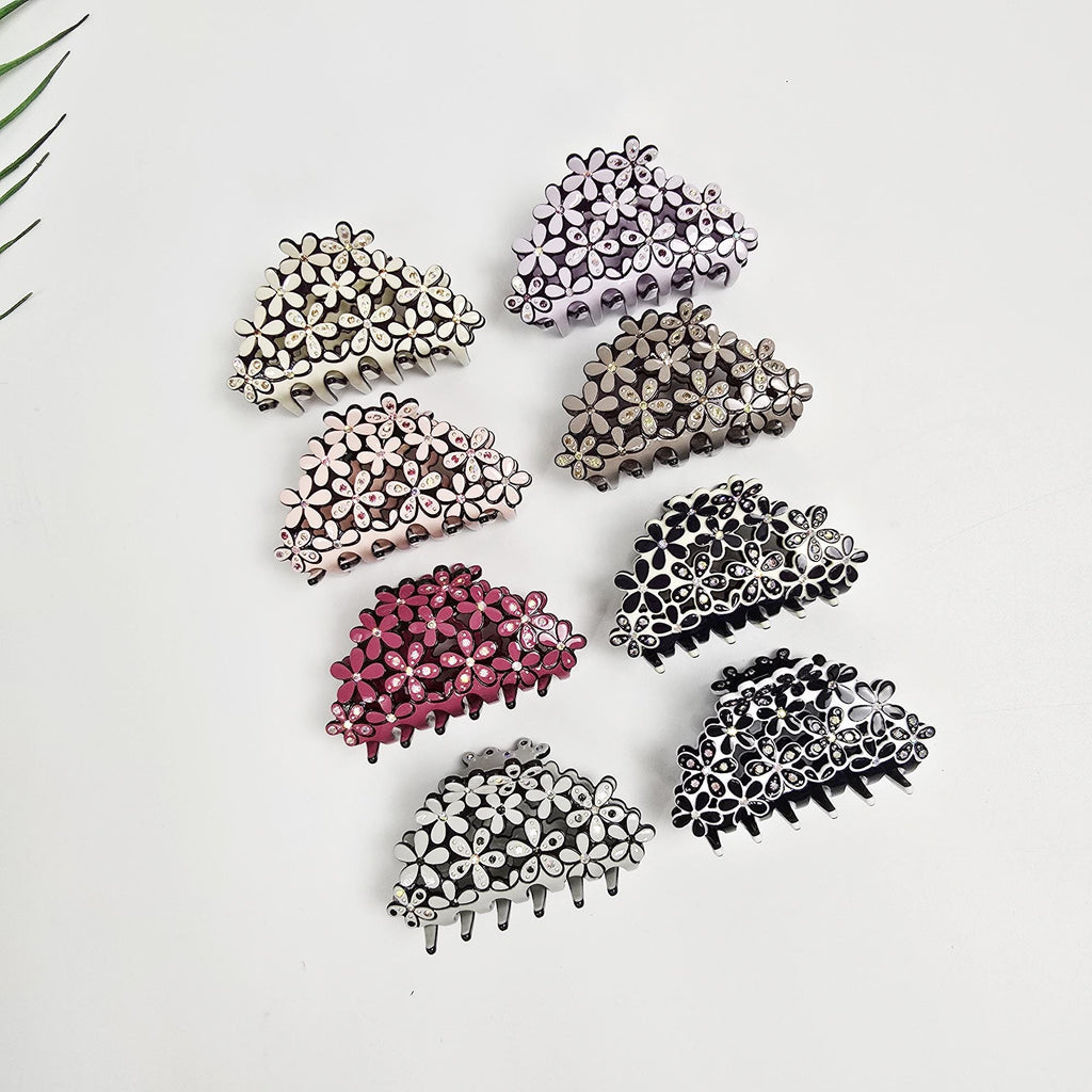 Cellulose acetate flower hair claw clip adorned with rhinestones. 2.6 inch small jaw clip available in pastel colors and black. Perfect for securing half-up hairstyles with a touch of sparkle. Assortment of eight floral hair claw clips in various colors: light purple, beige, pink, burgundy, dark brown, black, and white. Each clip features a half-moon shape adorned with small, flower-shaped embellishments. The clips are displayed against a white background.