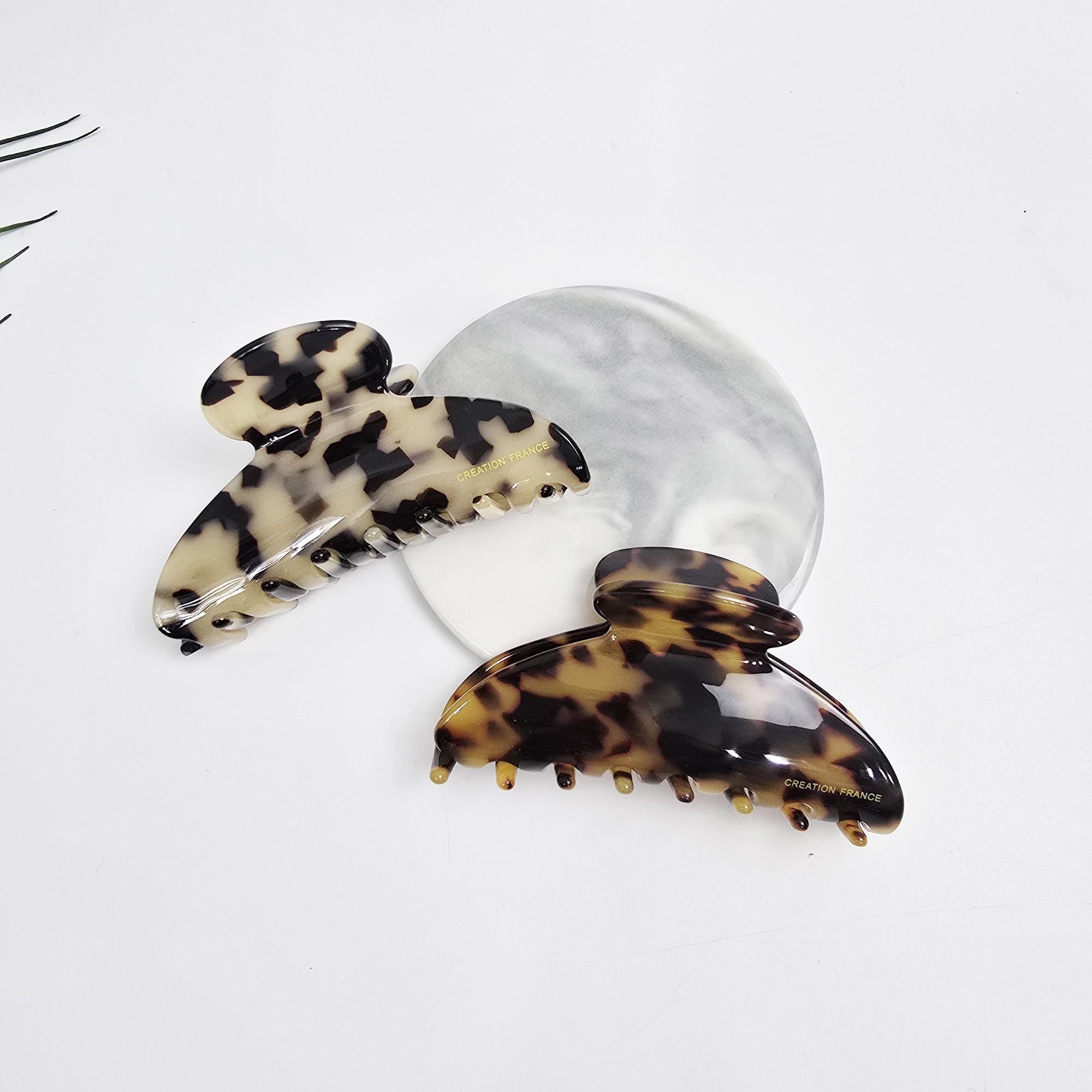 Cellulose acetate hair claw clip in half-moon shape featuring classic tortoise shell pattern. 3.5 inch medium jaw clip perfect for half-up hairstyles. Elegant French style accessory. Two tortoise shell hair clips. The clips are brown and black with a mottled pattern. The clips have a gold inscription that reads 'CREATION FRANCE'.