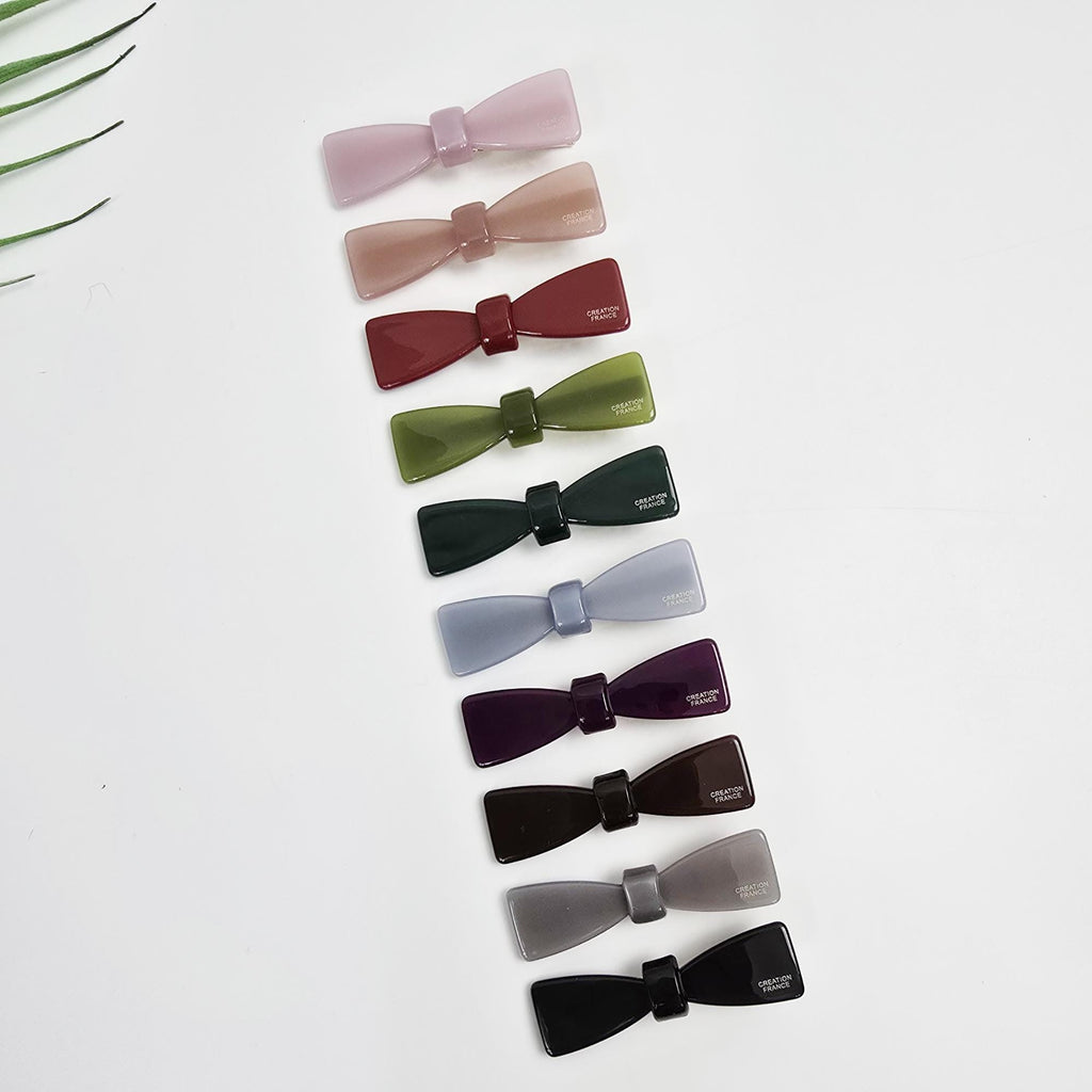Cellulose acetate bow hair clip in solid pastel colors. 2.4 inch alligator barrette for girls side bangs. Minimalist and cute hair accessory. Ten bow-shaped hair clips in various colors including pink, red, green, purple, black, and gray. Each clip has the words 'CREATIVE FRANCE' printed on the back.