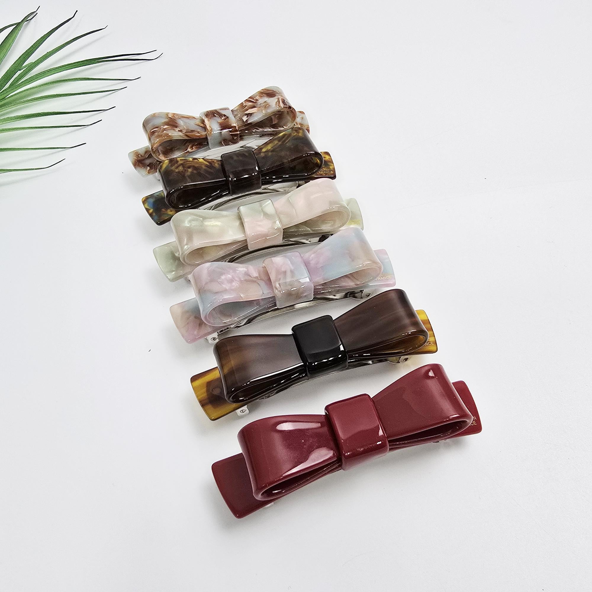 Cellulose acetate bow tie hair barrette in tortoise shell and pastel colors. 3.5 inch automatic french clip perfect for securing half-up hairstyles or low ponytails. Classic preppy hair accessory. A set of six hair clips with bow designs. The bows are made of different colors and patterns of plastic. The clips are silver and have a simple design.