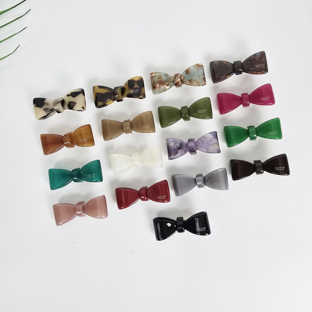 Cellulose acetate mini bow hair clip in green and tortoise shell patterns. Small non-slip alligator clip perfect for toddler girls or securing side bangs. Minimalist and cute hair accessory.  A collection of 17 assorted color bow hair clips. The hair clips are made of plastic and have a glossy finish. The colors include brown, black, green, purple, red, and white.