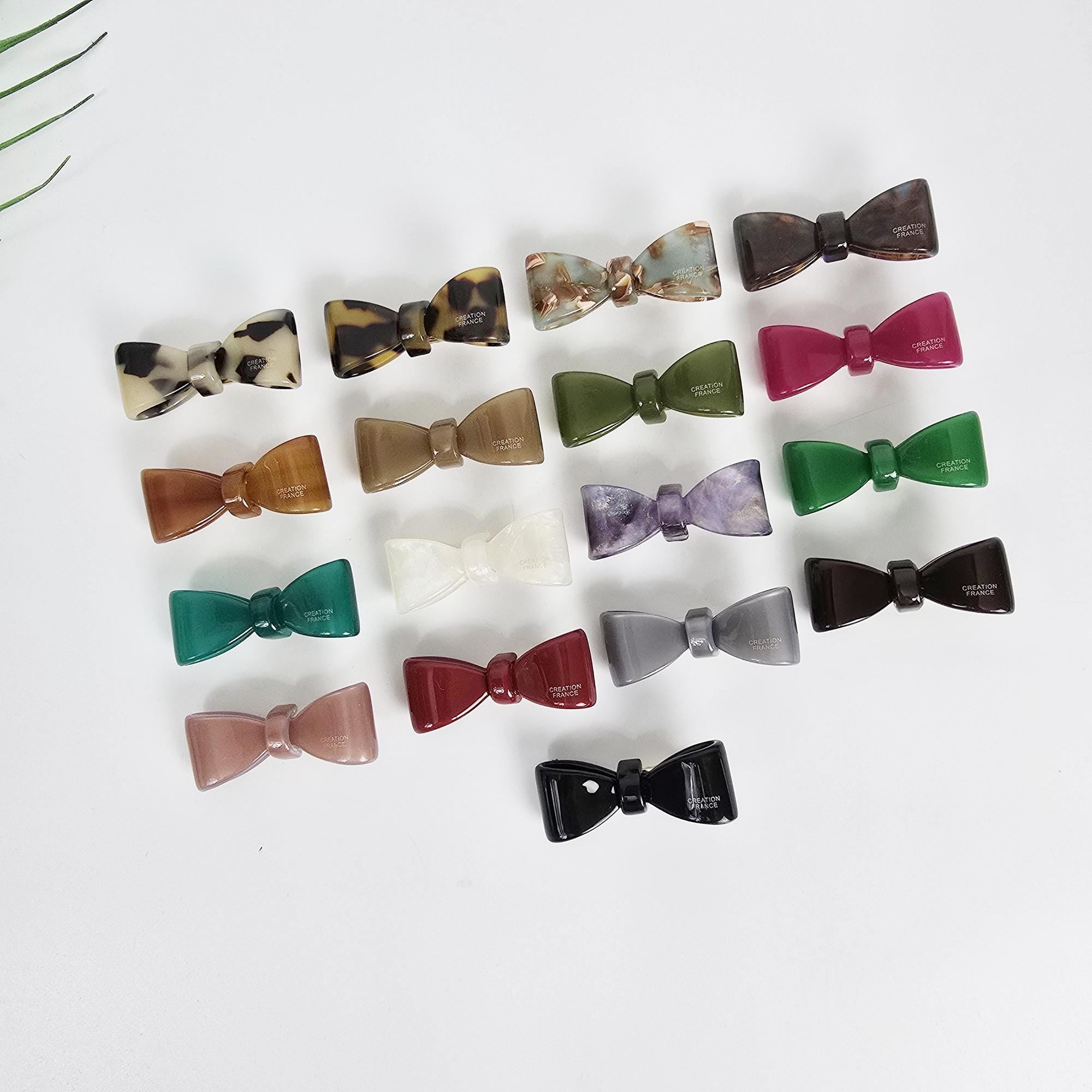 Cellulose acetate mini bow hair clip in green and tortoise shell patterns. Small non-slip alligator clip perfect for toddler girls or securing side bangs. Minimalist and cute hair accessory.  A collection of 17 assorted color bow hair clips. The hair clips are made of plastic and have a glossy finish. The colors include brown, black, green, purple, red, and white.