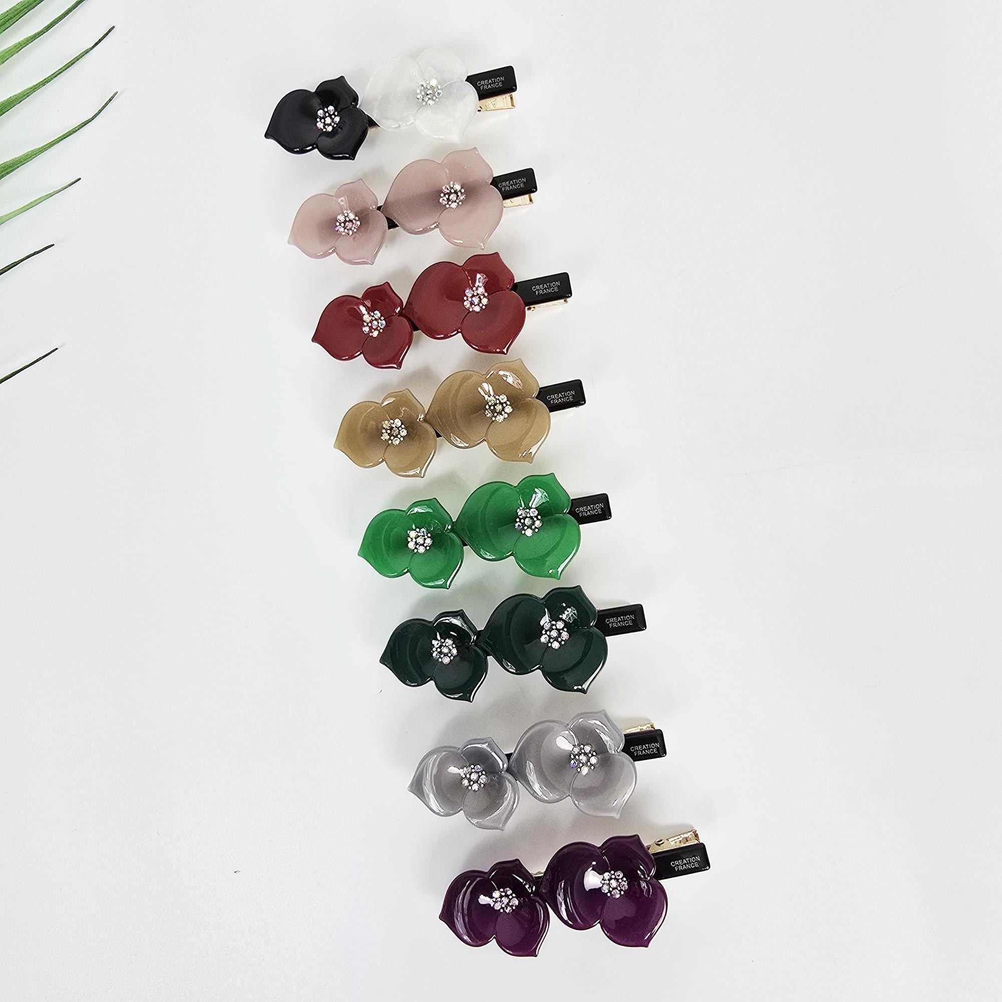 Cellulose acetate double flower hair clip with rhinestone center. 2.8 inch floral alligator barrette for women side hair and bangs. Eight pairs of hair clips with a flower design. The clips are in various colors including white, pink, red, beige, green, dark green, silver, and purple. Each clip has two flowers with a rhinestone in the center of each flower.