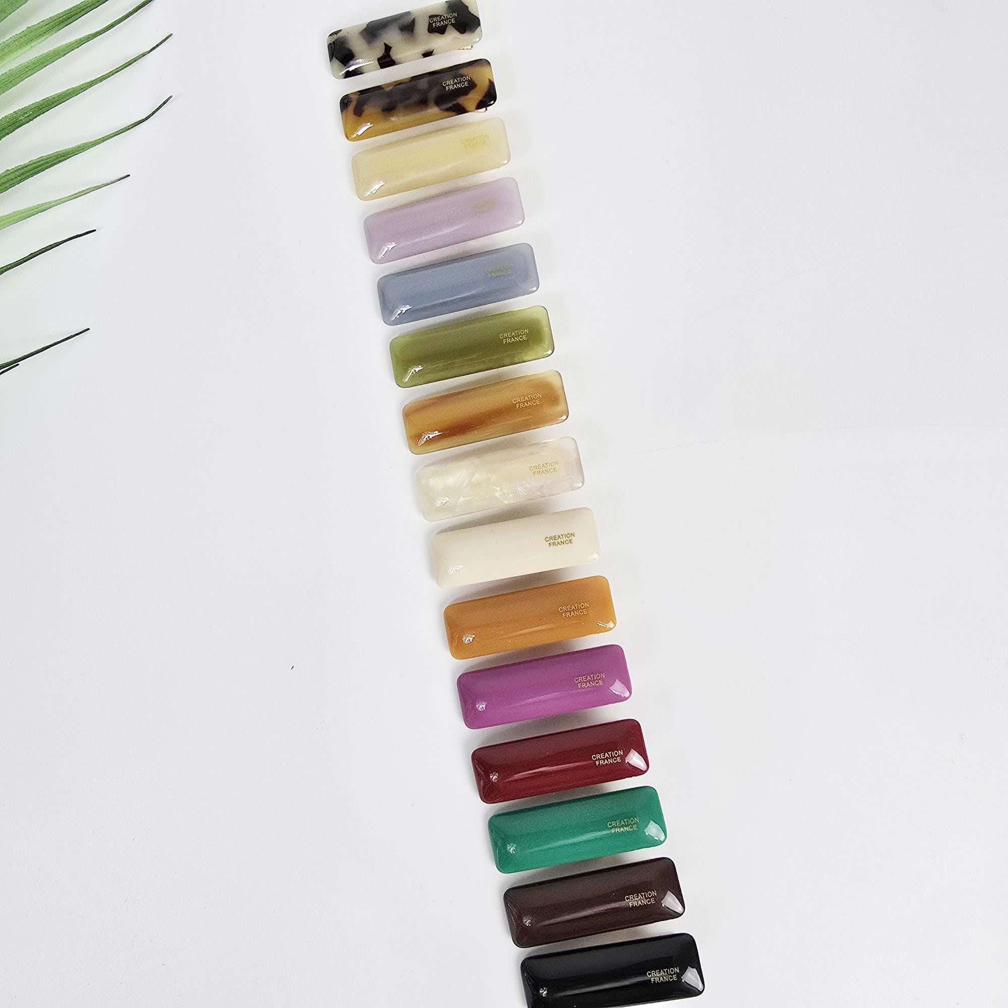 Cellulose acetate ultra mini hair clip in a simple convex bar shape. Small 1.9 inch solid and tortoise shell alligator barrette for toddlers and girls. A collection of 15 rectangular hair clips in various colors including brown, white, purple, blue, green, red, and black. The clips are arranged vertically in a row and have the words 'CREATOR PARIS' printed on them.