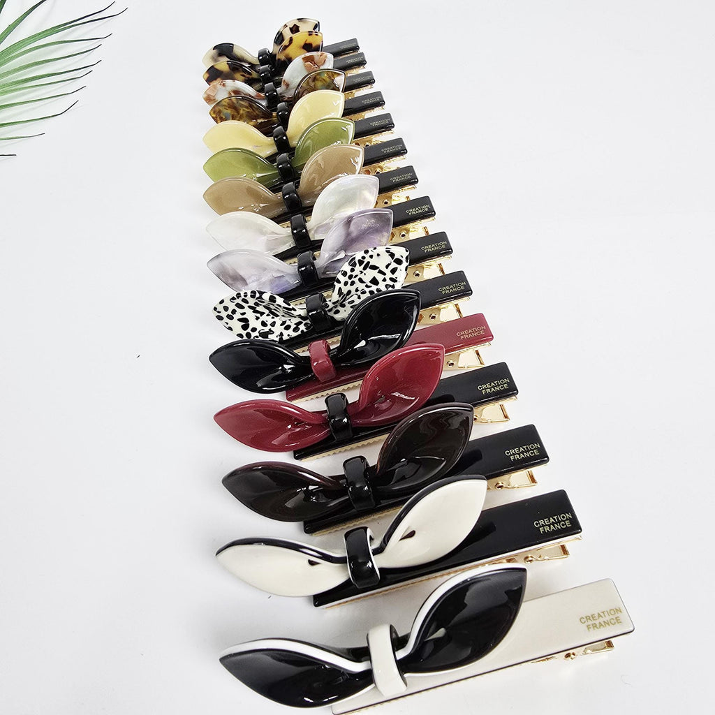 Cellulose acetate alligator hair clip featuring a unique lip-shaped bow design. 3.1 inch long duckbill barrette available in tortoise shell and solid colors. Perfect for holding side bangs. A collection of black and gold hair clips with various colored bows. The bows are made of different materials, including leopard print, white, red, and black. The clips are labeled 'Creation France'.