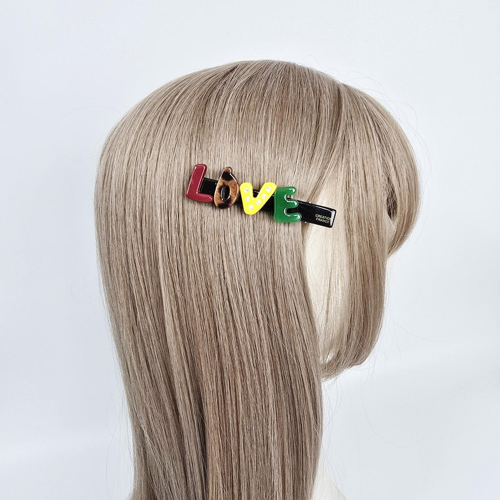 Cellulose acetate LOVE word hair clip with colorful letters. 3 inch alphabet side barrette with tortoise shell accent for girls and women. A black hair clip with the word 'LOVE' spelled out in colorful letters. The letters are red, brown with black spots, yellow with white dots, green, and black. The letters are attached to a black clip with the words 'CREATIVE FRANCE' printed on the back.