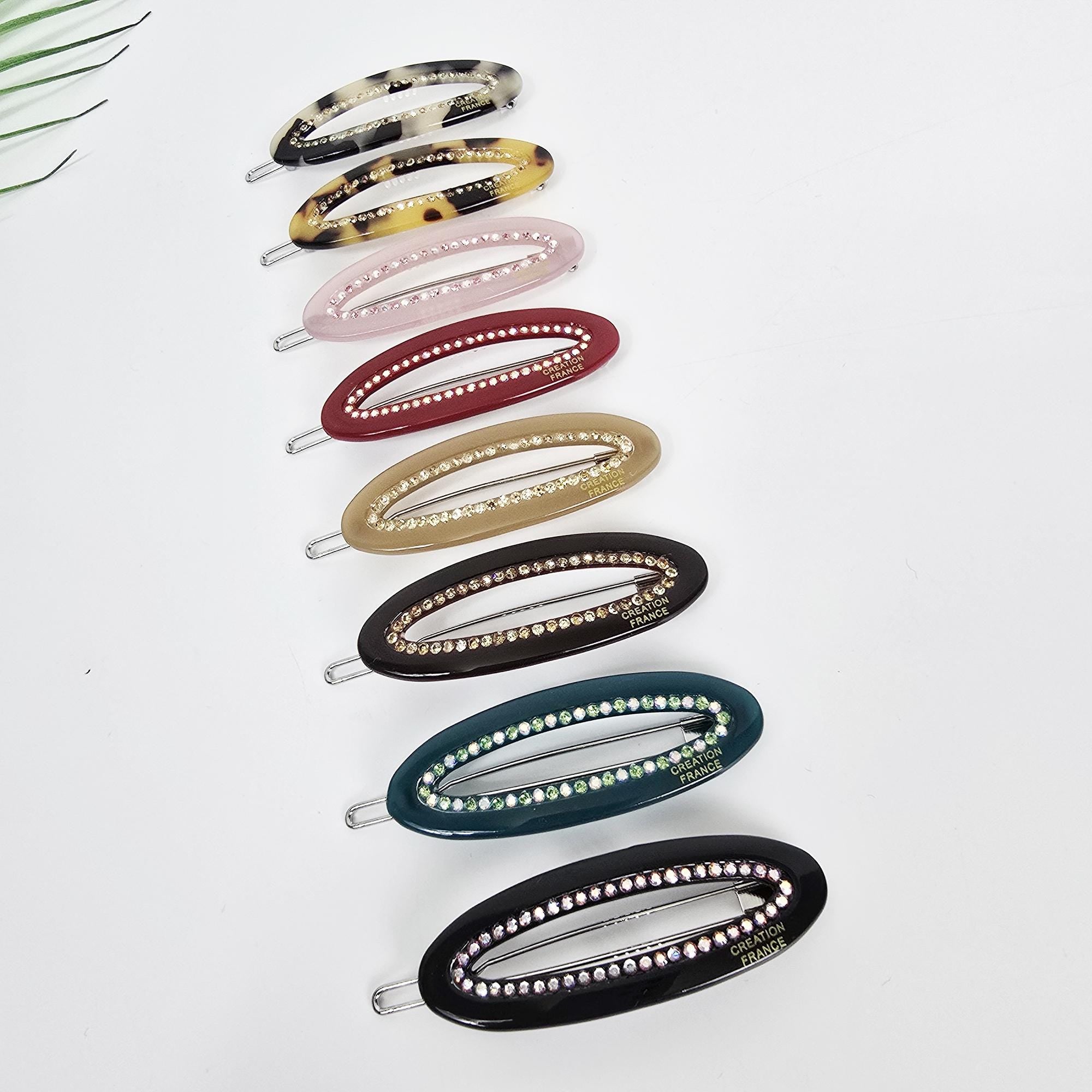 Cellulose acetate oval snap hair clip with rhinestone border. 2.3 inch tortoise shell and pink sparkly barrette for bangs and side hair. A set of eight oval-shaped hair clips with rhinestones. The clips are in various colors including brown, beige, red, green, and black. Each clip has the words 'CREATION FRANCE' printed on it.