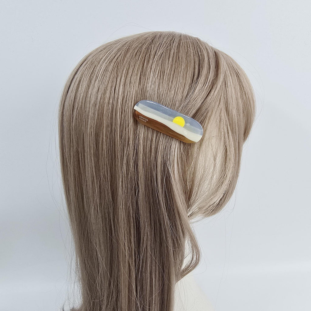 Cellulose acetate hair clip featuring a sunset landscape design with sun and hills. 2.5 inch side alligator barrette for women and girls. A gray, white, and yellow hair clip with a sun design. The clip is made of resin and has a shiny finish.