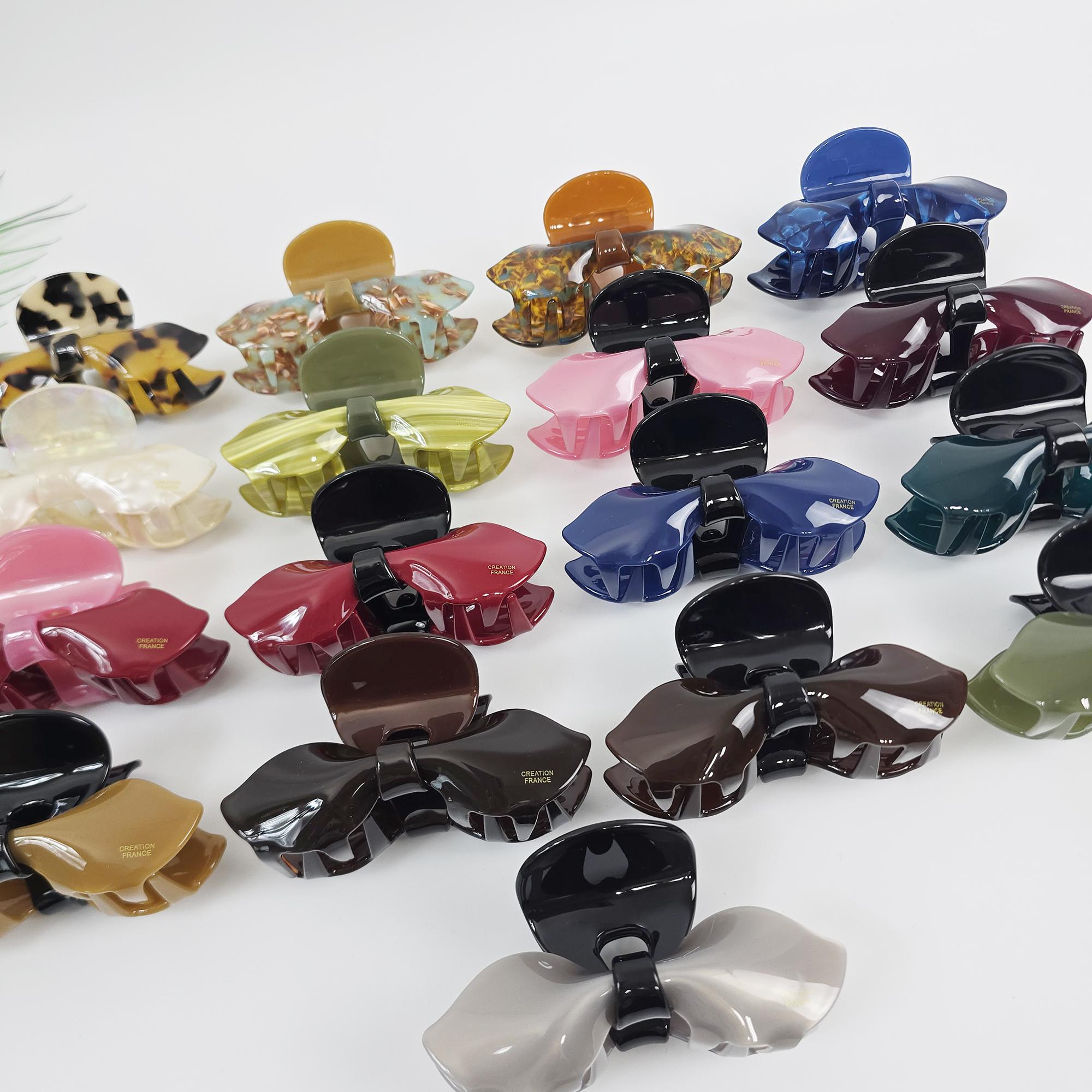 Cellulose acetate ribbon hair claw clip in pink and tortoise shell. Large 3.9 inch bow design jaw clamp for women updos and French twists. A collection of 18 assorted plastic hair clips in various colors including brown, pink, green, blue, black, and white. The clips are shaped like a bow and have a textured surface. The clips are made by the brand 'Accessorize France'.