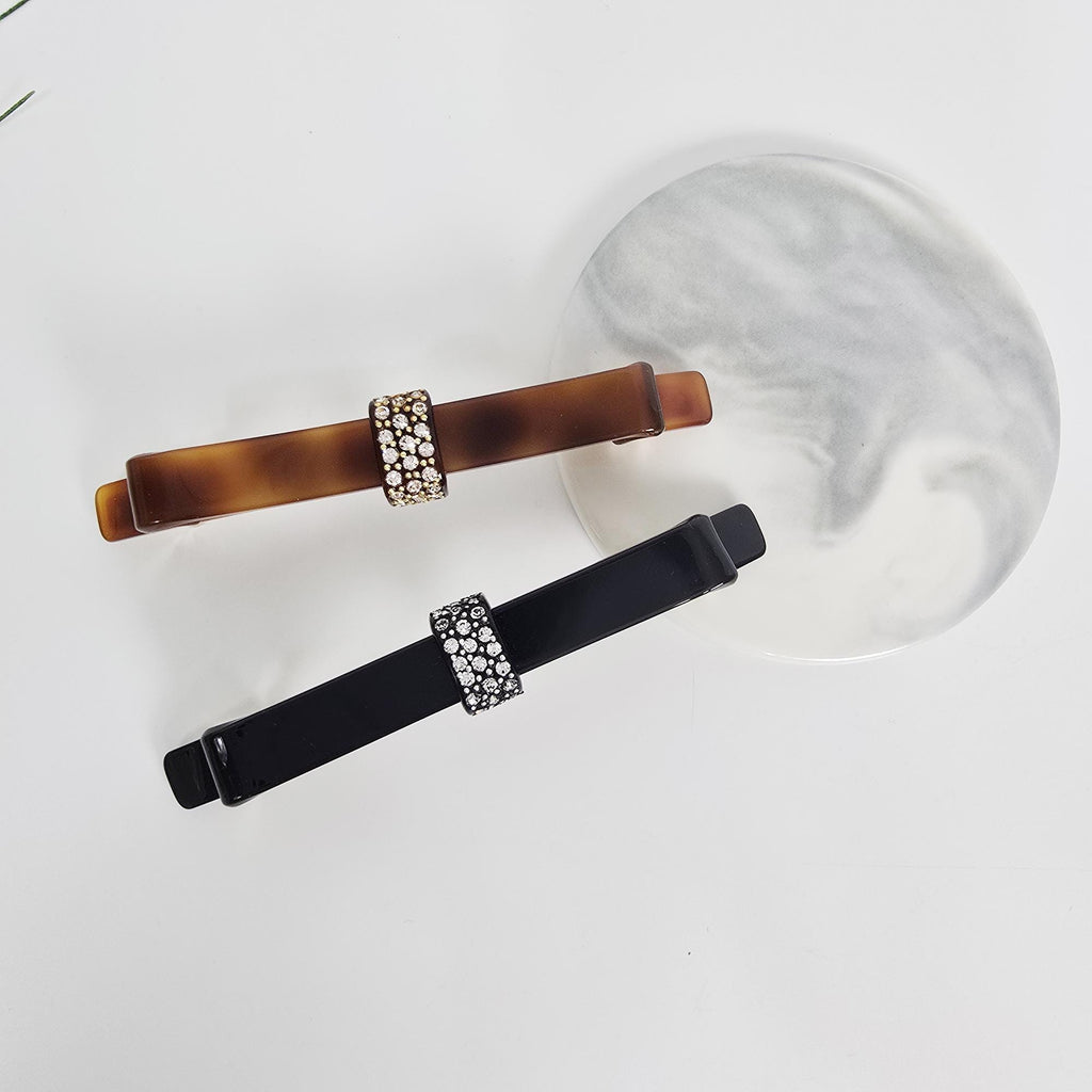 Cellulose acetate long bow barrette with crystal accents in tortoise shell and black for elegant ponytail styling.Two barrettes with rhinestone accents. One is brown with a tortoiseshell pattern, and the other is black. Both have a rectangular shape with a band of rhinestones in the center. They are displayed on a white surface with a decorative, round, marble-patterned plate.