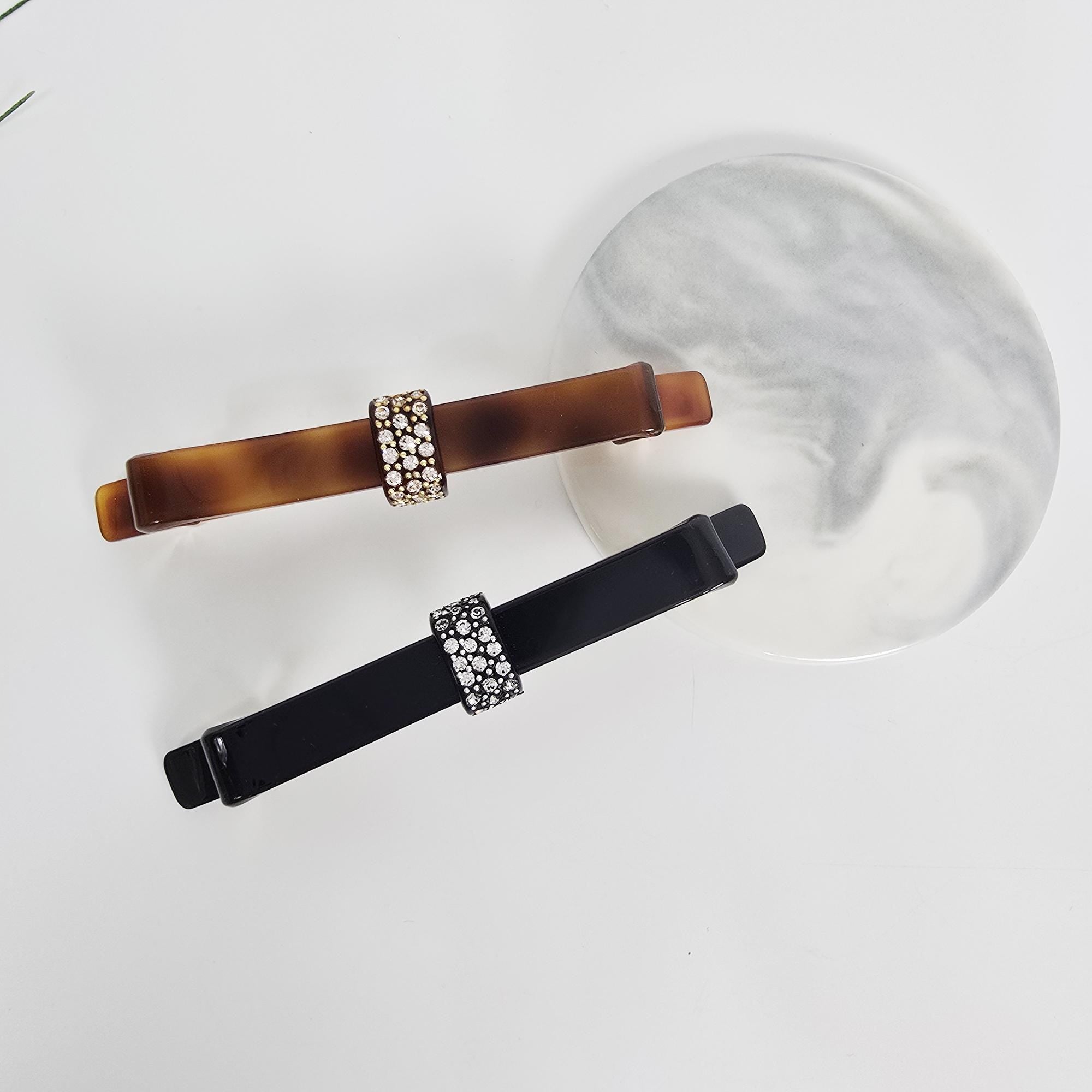 Cellulose acetate long bow barrette with crystal accents in tortoise shell and black for elegant ponytail styling.Two barrettes with rhinestone accents. One is brown with a tortoiseshell pattern, and the other is black. Both have a rectangular shape with a band of rhinestones in the center. They are displayed on a white surface with a decorative, round, marble-patterned plate.