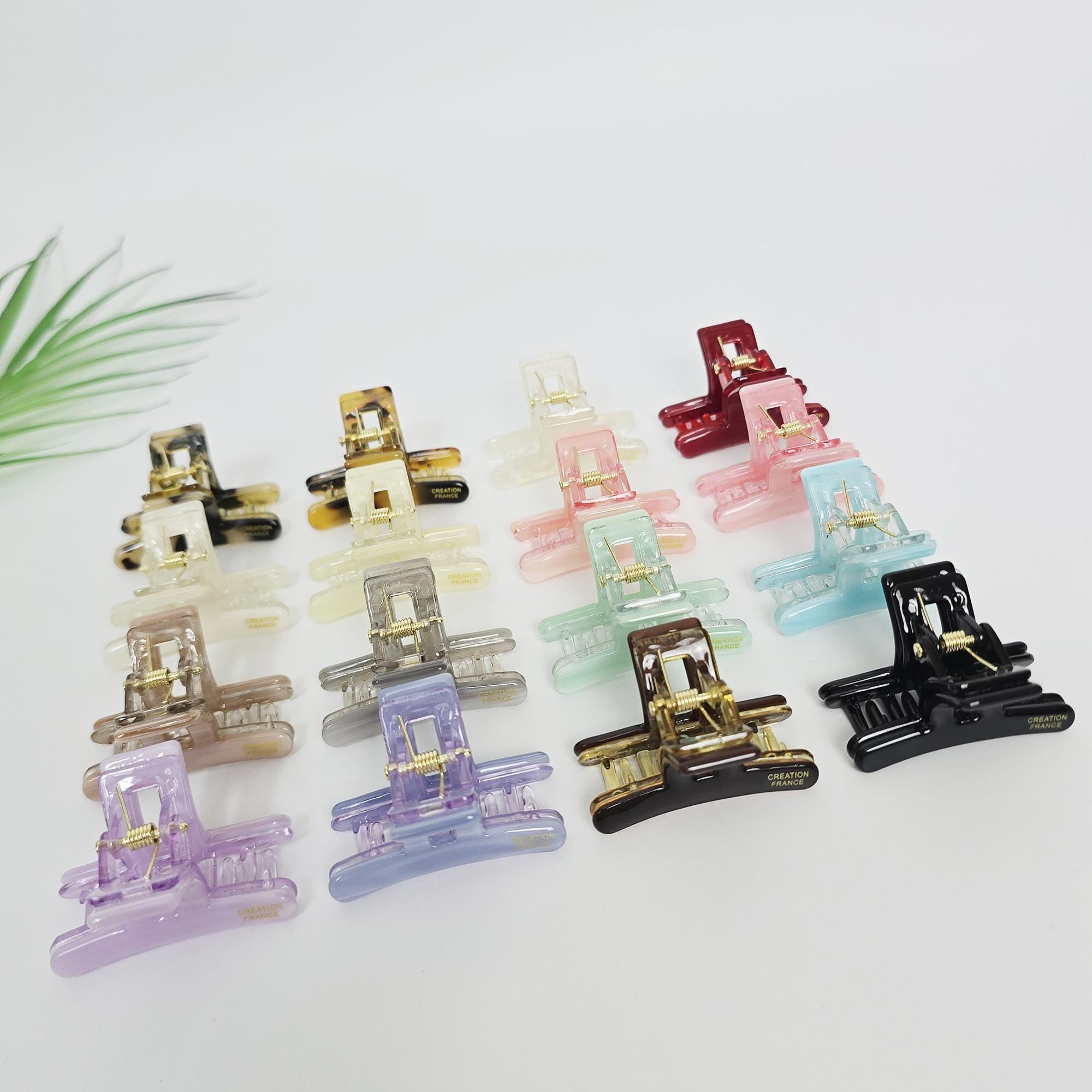 Cellulose acetate rectangular mini hair claw clip with geometric cutout design in pastel and tortoise colors for half-up styling. A collection of small, colorful hair clips. The clips are in various shades including black, brown, clear, gold, pink, red, green, blue, purple, and gray. Each clip has a rectangular shape with a hinge and a decorative element. The clips are arranged on a white surface.