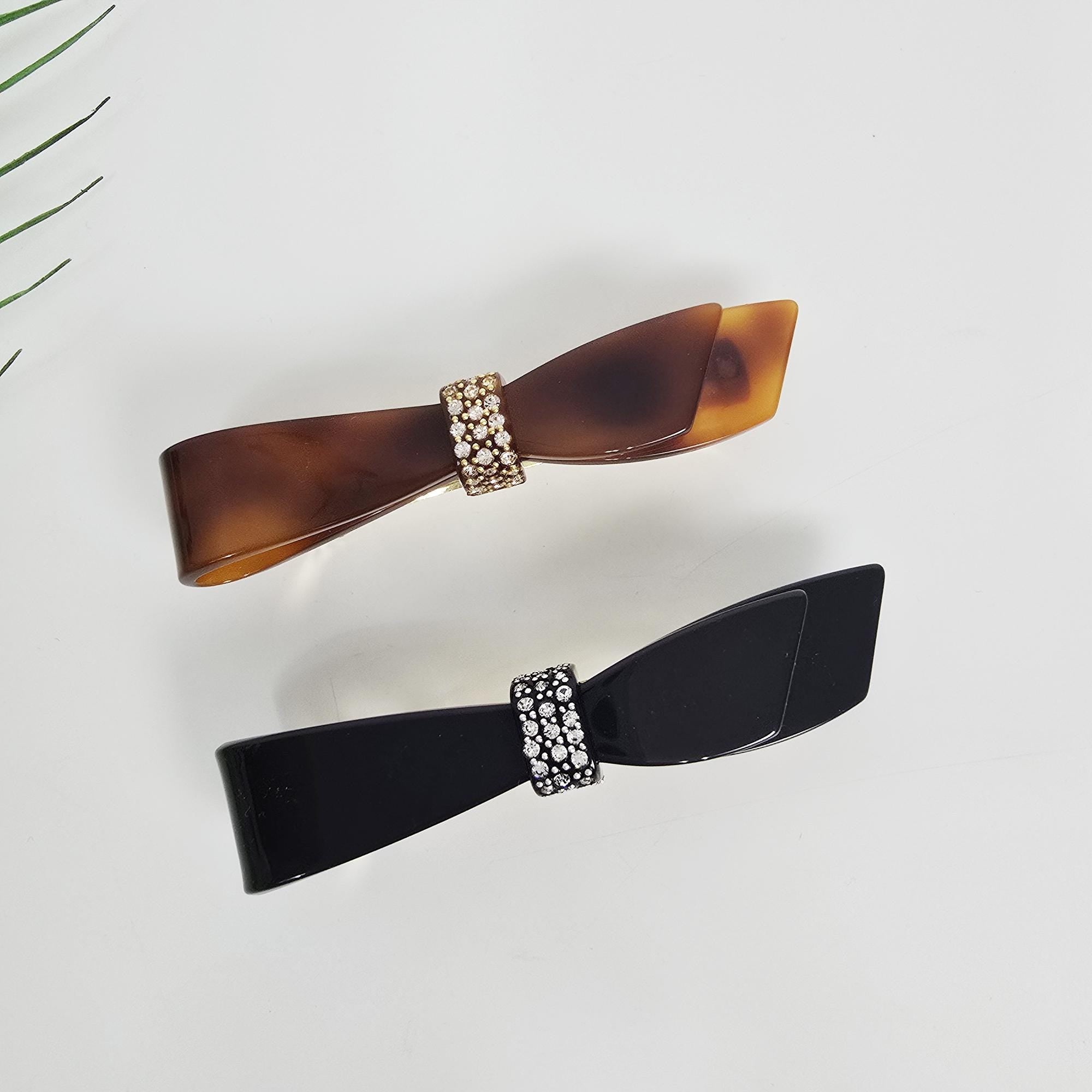 Cellulose acetate single knot bow barrette with Austrian crystal accents in tortoise shell and black for elegant ponytail styling. Two bow-shaped hair clips. One clip is a tortoiseshell pattern with a gold-colored rhinestone center. The other clip is black with a silver-colored rhinestone center. Both clips are on a white background.