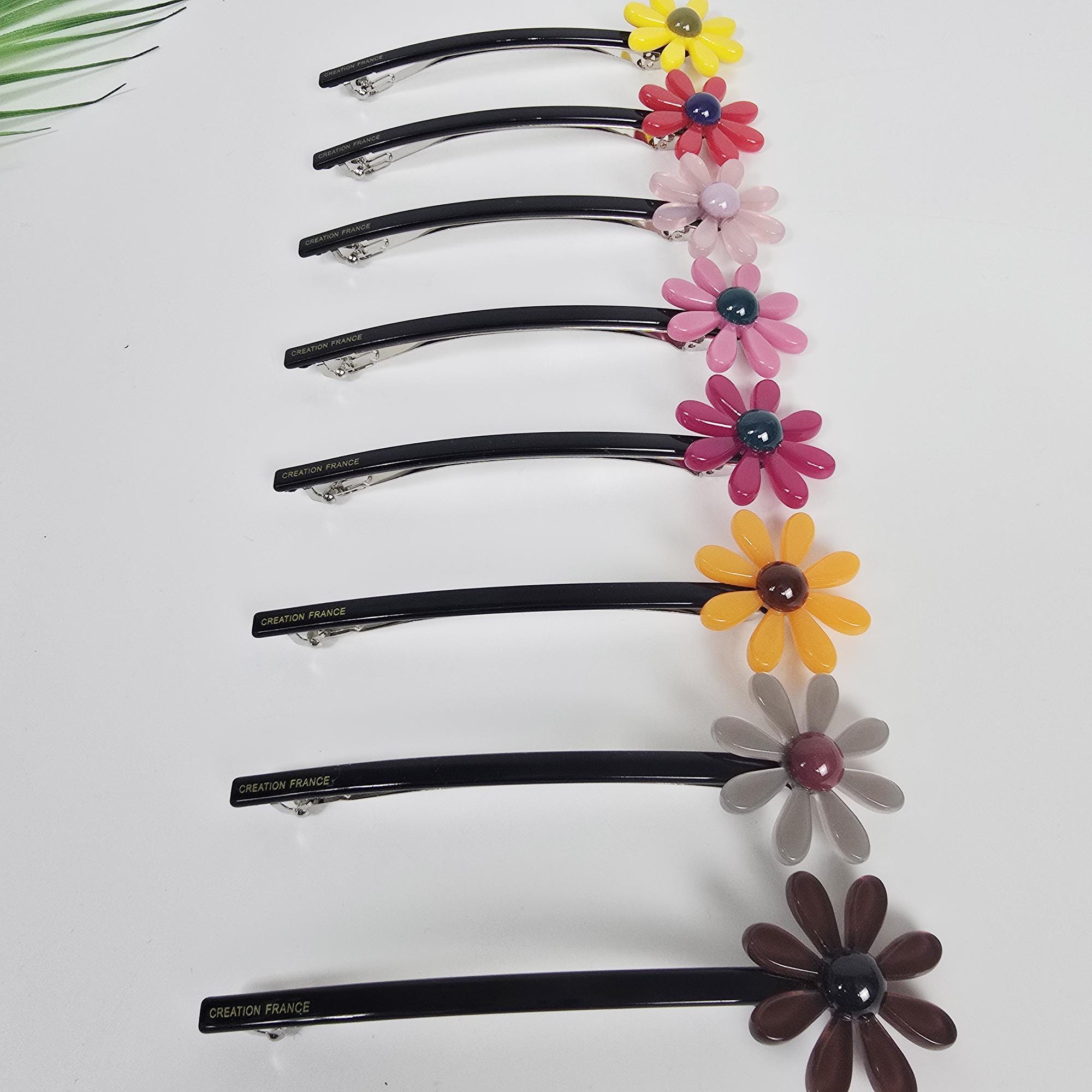 Cellulose acetate slim long hair barrette with sunflower design for ponytail holder and half-up styling. An assortment of daisy flower hair clips in various colors. The clips have black bases with a curved design and metal clasps. The daisy flowers are yellow, red, pink, purple, orange, gray, and brown. The text 'CREATION FRANCE' is visible on some clips.