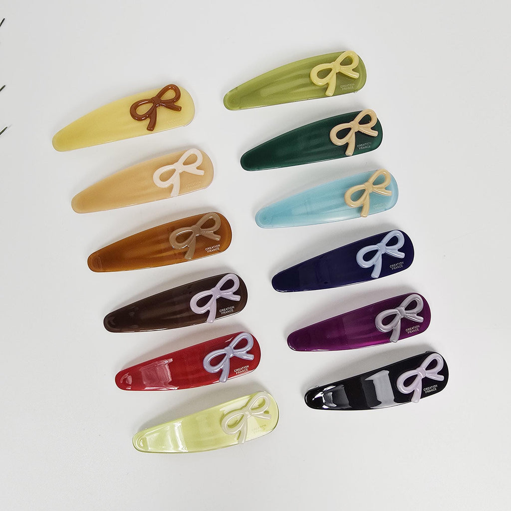 Cellulose acetate triangle snap hair clips with mini bow detail in various colors for side bangs styling. A collection of colorful hair clips with bow accents. The clips are in various colors including yellow, green, blue, navy, purple, red, and black. Each clip has a bow in a contrasting color, and some have text that reads 'CREATION FRANCE'.