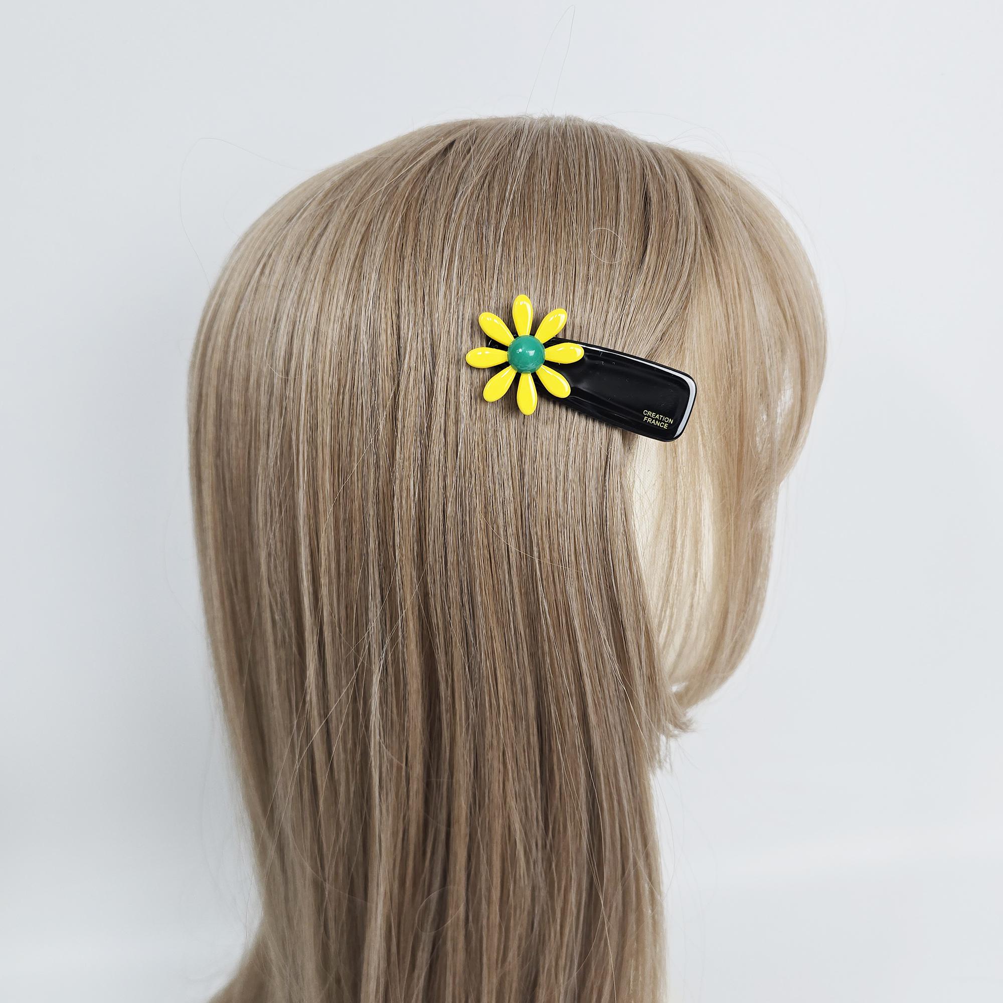 Glossy acetate daisy hair clip in colorful floral shape with black base for side bangs and girls hair styling featuring smooth edges and secure grip. A black hair clip with a yellow daisy flower and a green center. The clip is attached to light brown hair. The flower is on the left side of the clip. The clip has the words 'CREATION FRANCE' printed on it.