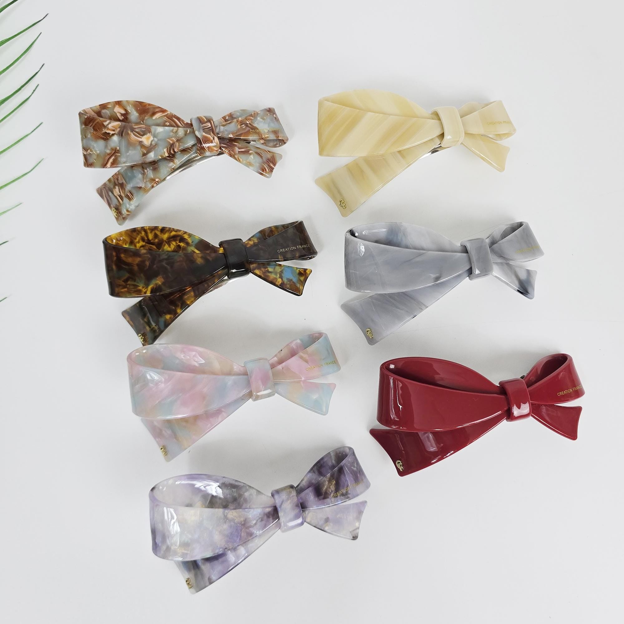 Cellulose acetate large bow barrette featuring a curved shape and french clip mechanism. Glossy red and tortoise shell patterns for thick hair or half-up styles. Collection of bow-shaped hair clips in various colors. The clips include shades of brown, yellow, gray, pink, and red. Each clip features a bow design with a clasp. The clips are made of a glossy material.
