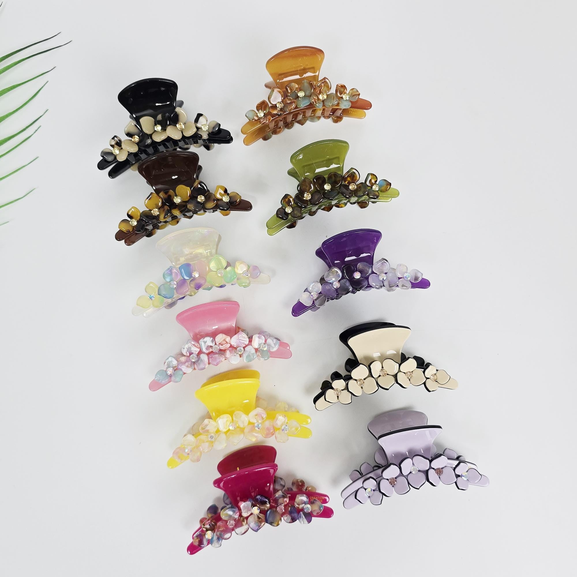 Cellulose acetate floral hair claw clip with rhinestone accents and tortoise shell pattern for women styling thick hair. A collection of hair claw clips in various colors. The clips are adorned with flower-shaped embellishments. Colors include black, brown, orange, green, purple, pink, yellow, and lavender. The clips are arranged on a white surface.
