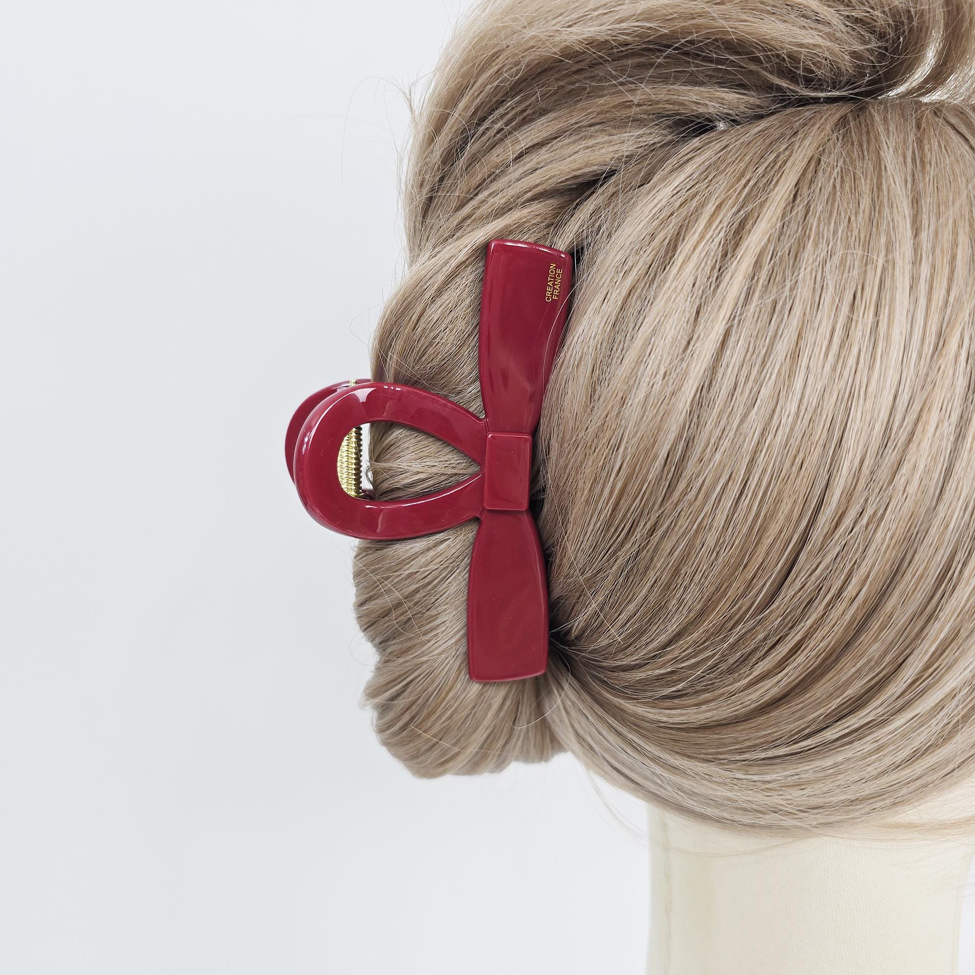 Cellulose acetate bow tie hair claw clip in tortoise shell and marble colors for elegant updos. A burgundy hair claw clip in a bow shape. The clip is made of glossy plastic and features a curved design with a rectangular center. The clip is holding a section of blonde hair, showcasing its functionality as a hair accessory. The clip has the words 'CREATION FRANCE' printed on it.