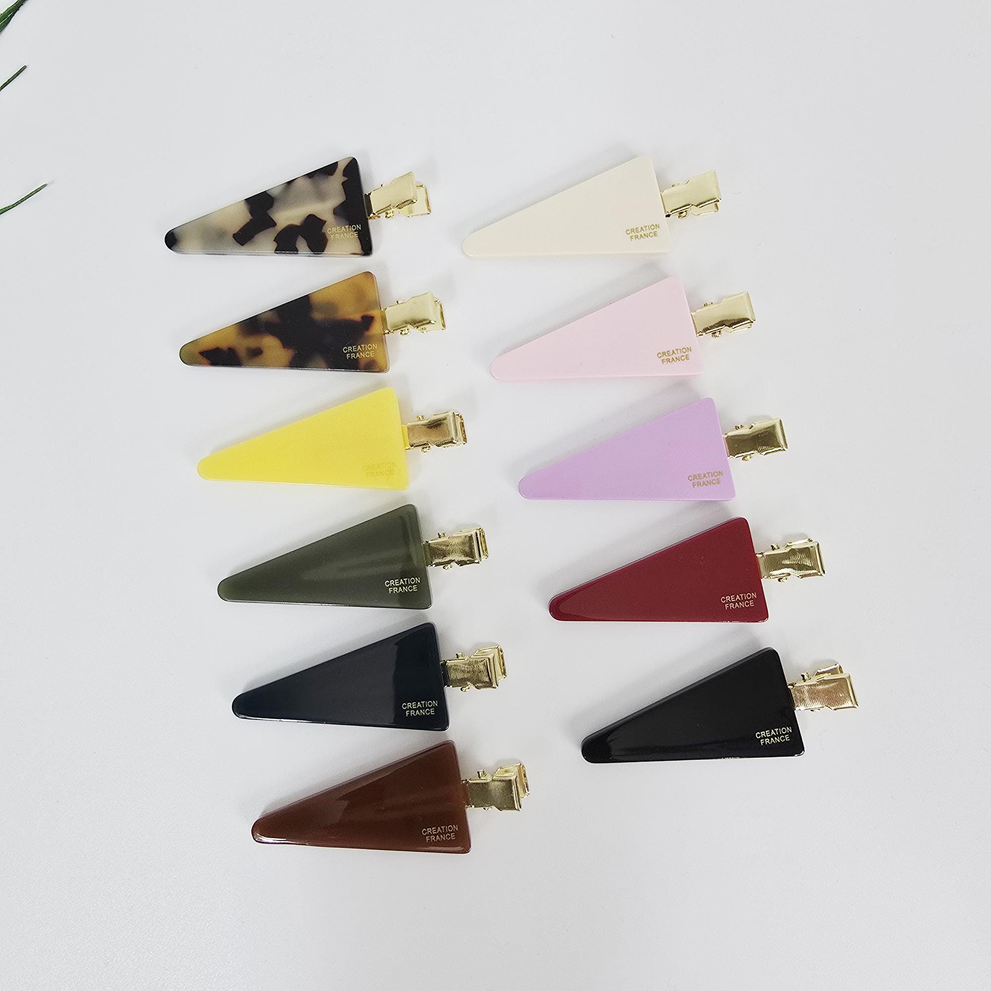 Cellulose acetate triangle flat hair clips in tortoise shell and pastel colors for creaseless makeup styling. A collection of triangular hair clips in various colors. The clips include patterns like tortoiseshell, and solid colors such as yellow, olive green, navy, and brown. Each clip has a gold-colored metal clasp and the text 'CREATION FRANCE'.